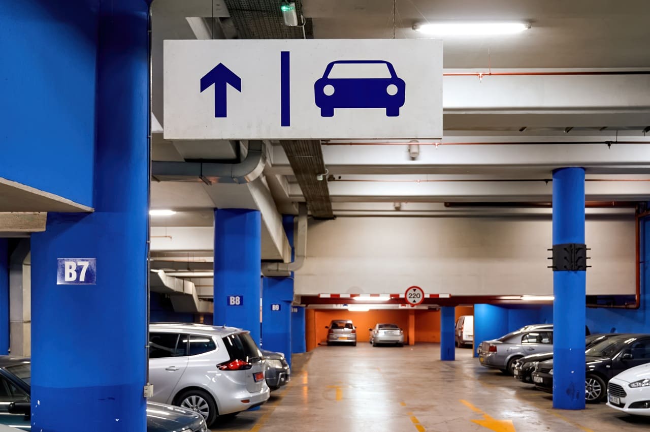 Interior de um estacionamento de prédio coberto, com vários carros estacionados e uma placa suspensa indicando direção.