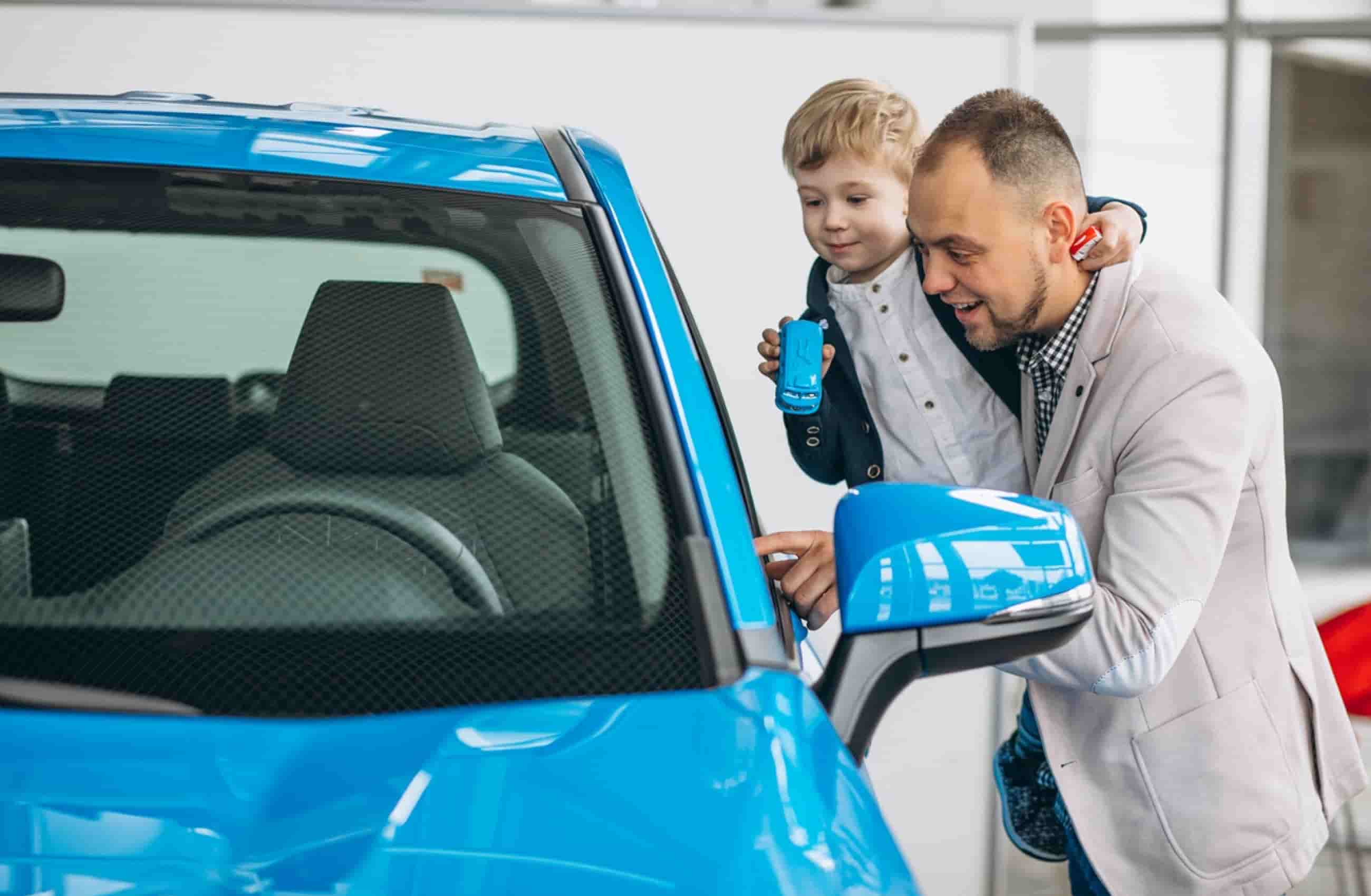 Homem branco com terno cinza segura um menino ainda criança loiro e banca no colo. Ambos estão analisando um carro azul na concessionária. A imagem representa carros que custam menos de 50 mil reais.
