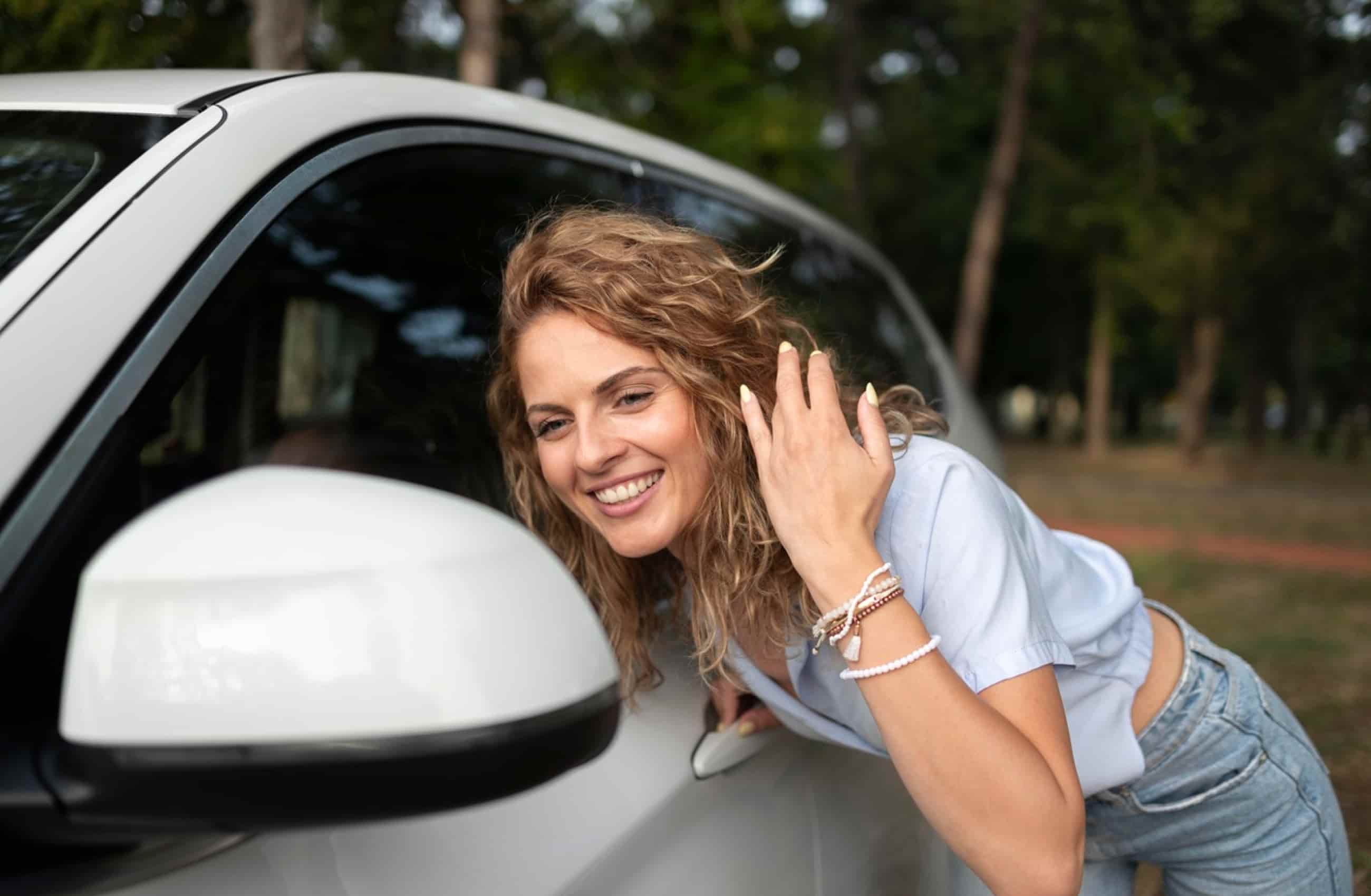 Mulher de camiseta branca e calça jeans está com as mãos no cabelo esvoaçante, enquanto se apoia no carro para olhar no retrovisor. A imagem representa carros que custam menos de 50 mil reais.