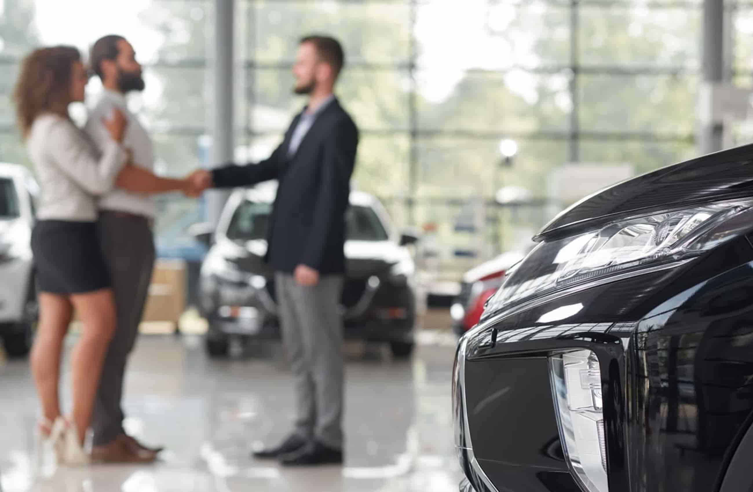 Um casal está cumprimentando um homem vestido com roupa social, ambos estão em segundo plano. Em primeiro plano temos a frente de um carro preto. A imagem representa a compra de um novo carro.