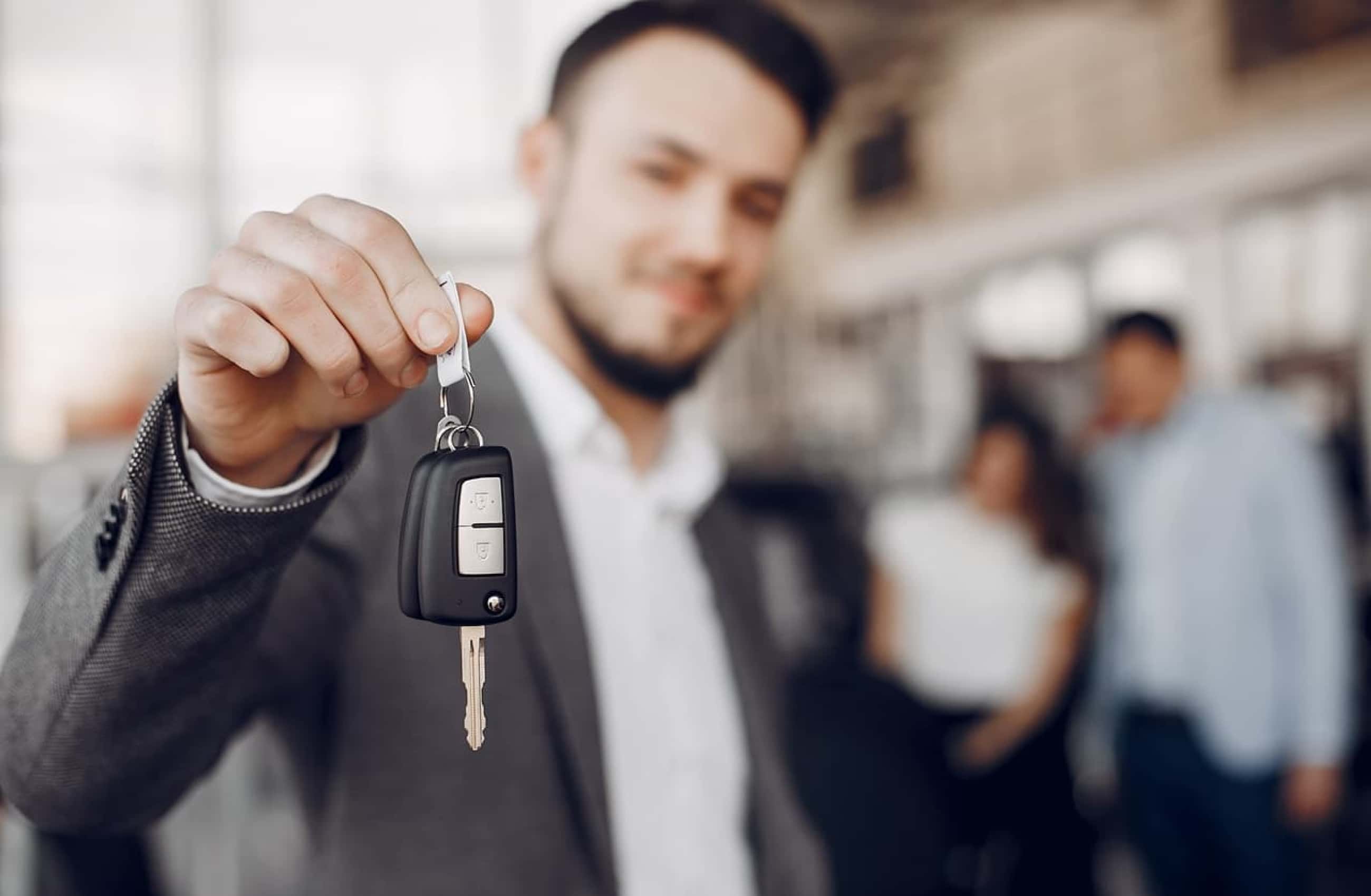 Homem branco vestido com terno cinza está em segundo plano na imagem. Em primeiro plano aparece uma chave de carro que ele está segurando. A imagem representa a importância de regularizar o carro alienado.