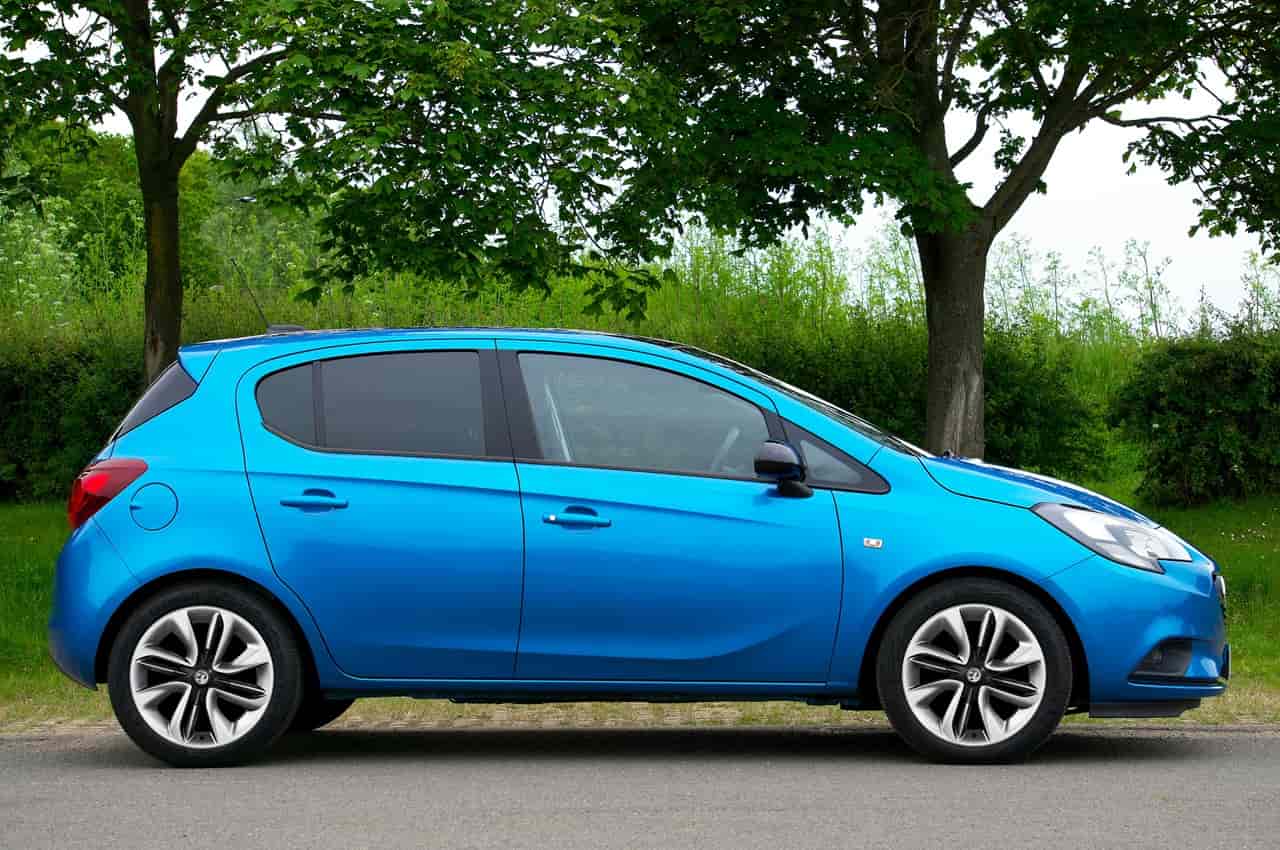 Carro hatch azul claro estacionado de perfil em uma rua com árvores e vegetação verde ao fundo.