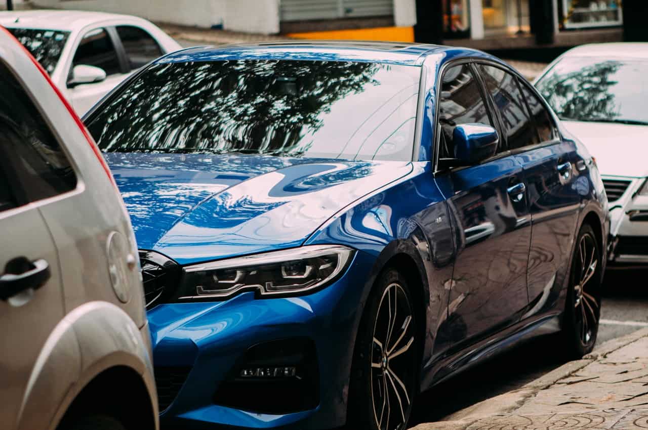 Carro sedan azul brilhante estacionado em uma rua urbana com reflexos de árvores na lataria.
