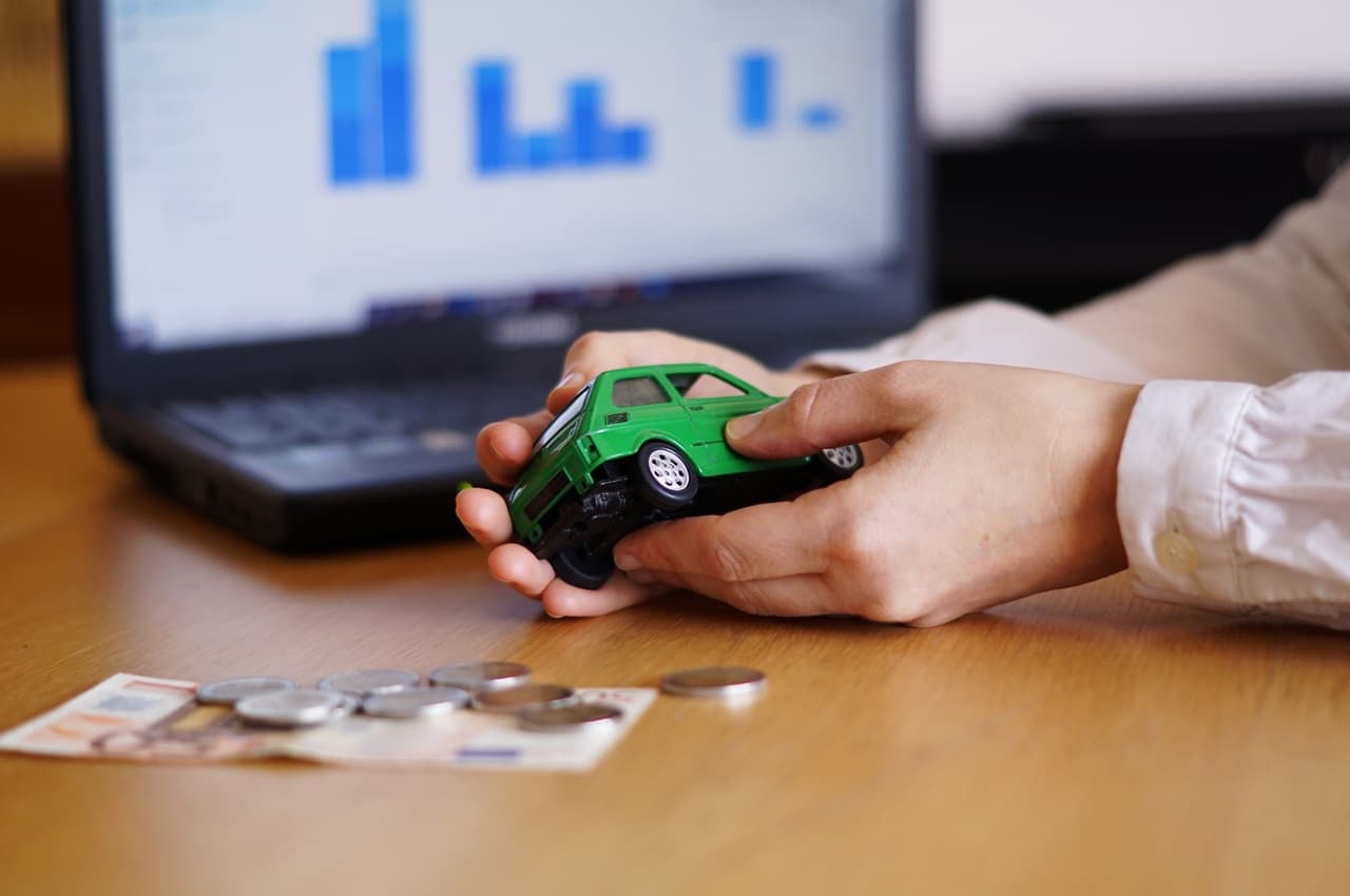 Mãos segurando uma miniatura de um carro verde sobre uma mesa de madeira, com moedas e uma nota de dinheiro, simbolizando um carro econômico.