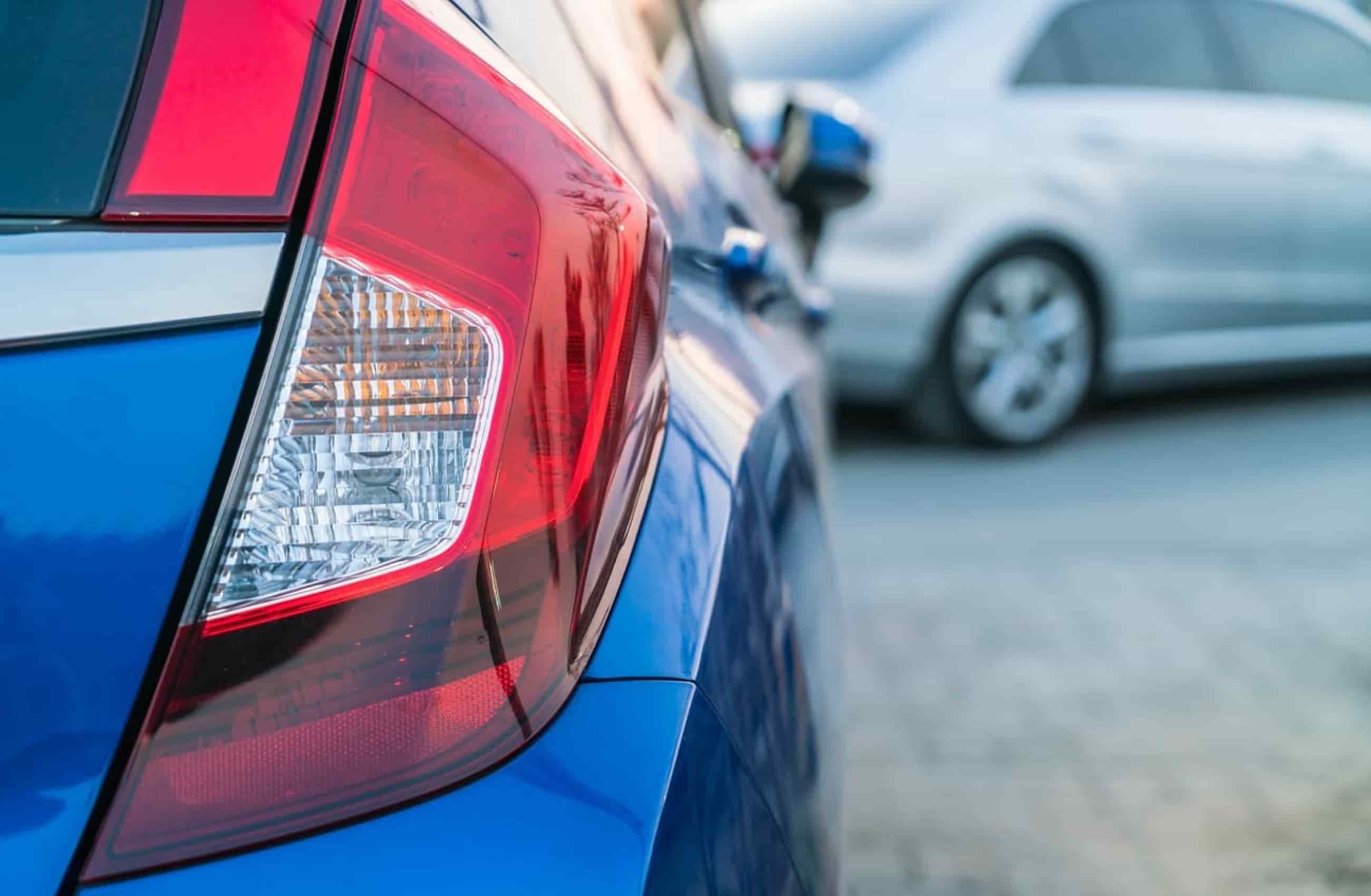 Lanterna traseira de um carro azul em foco. Ao fundo, um carro prata desfocado aparece a frente do carro azul.