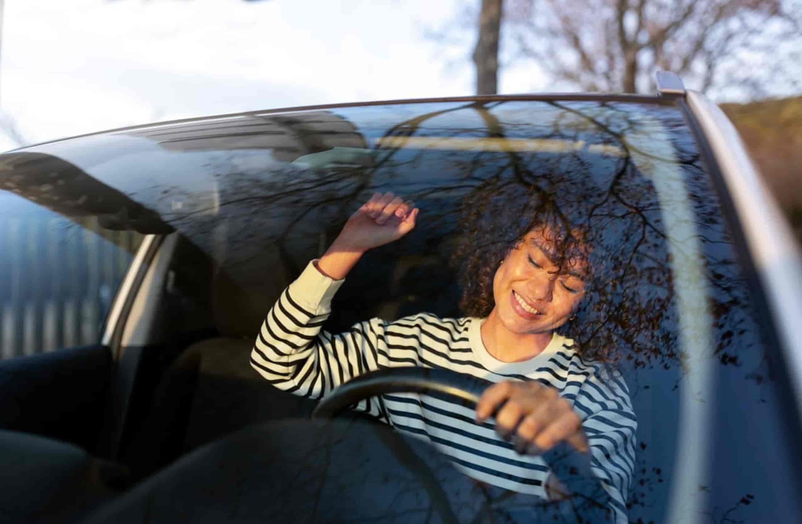 Mulher vestindo um casaco listrado preto e branco sorri enquanto dança dentro do carro, com uma das mãos erguidas e outra no volante.