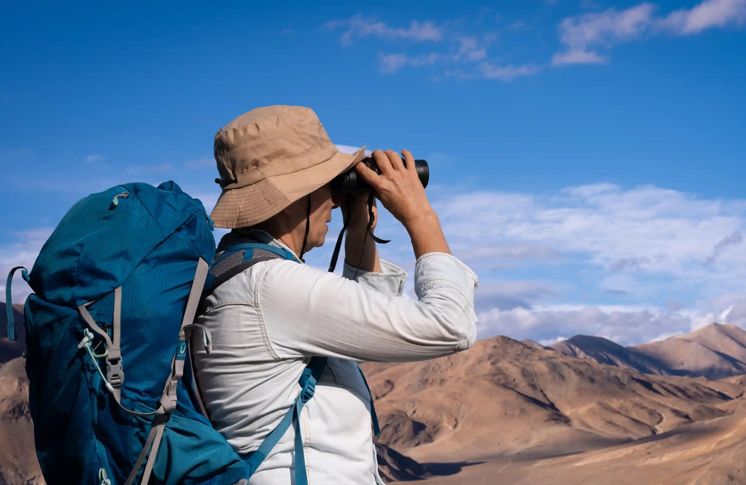 Pessoa de perfil, usando chapéu e mochila grande, observando a paisagem montanhosa com binóculos em dia claro.