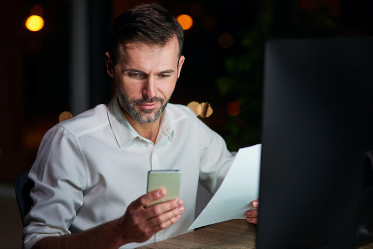 Homem consultando o celular enquanto analisa um documento em frente ao computador, em contexto de organização financeira e investimentos.