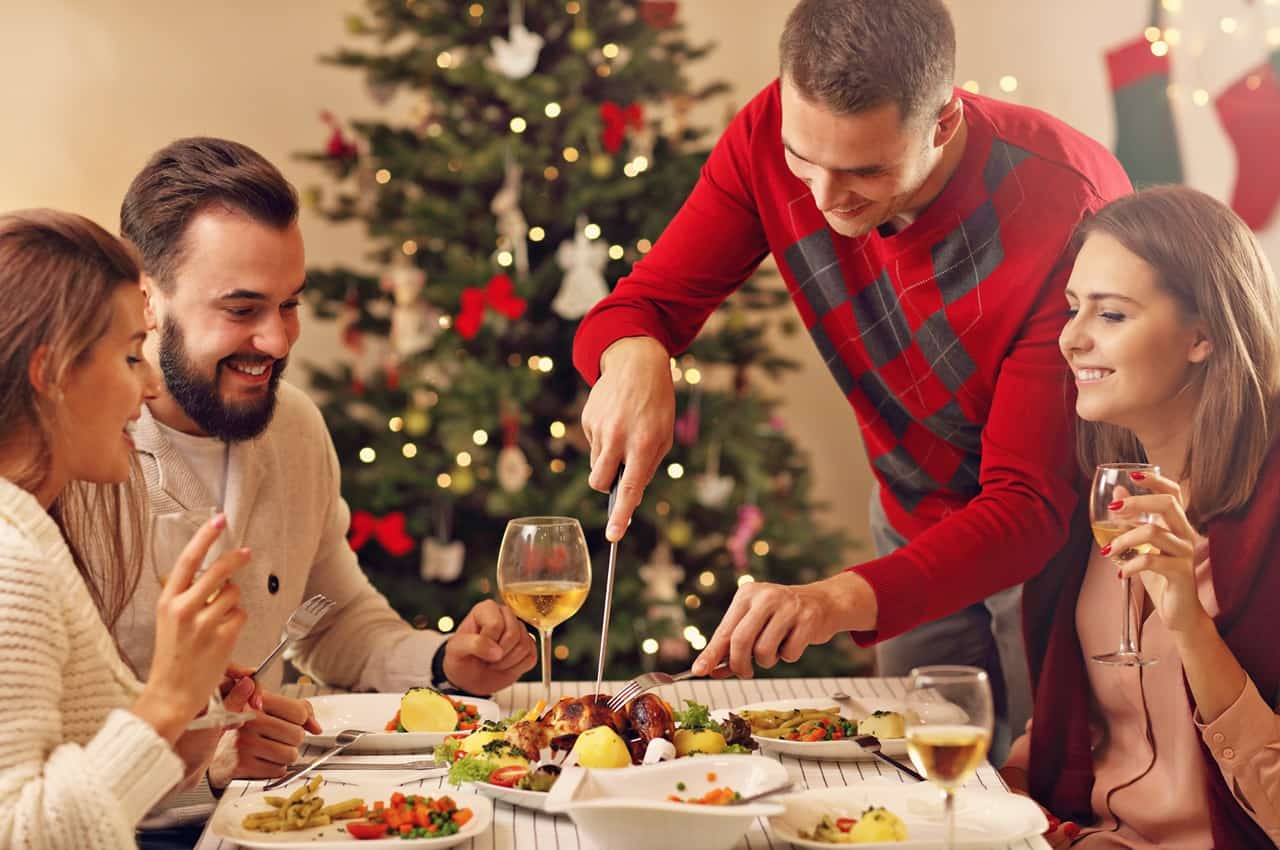 Grupo de pessoas sorridentes (duas mulheres e dois homens) realizando a ceia de natal. O homem corta um peru com garfo e faca, enquanto as pessoas bebem champanhe. 