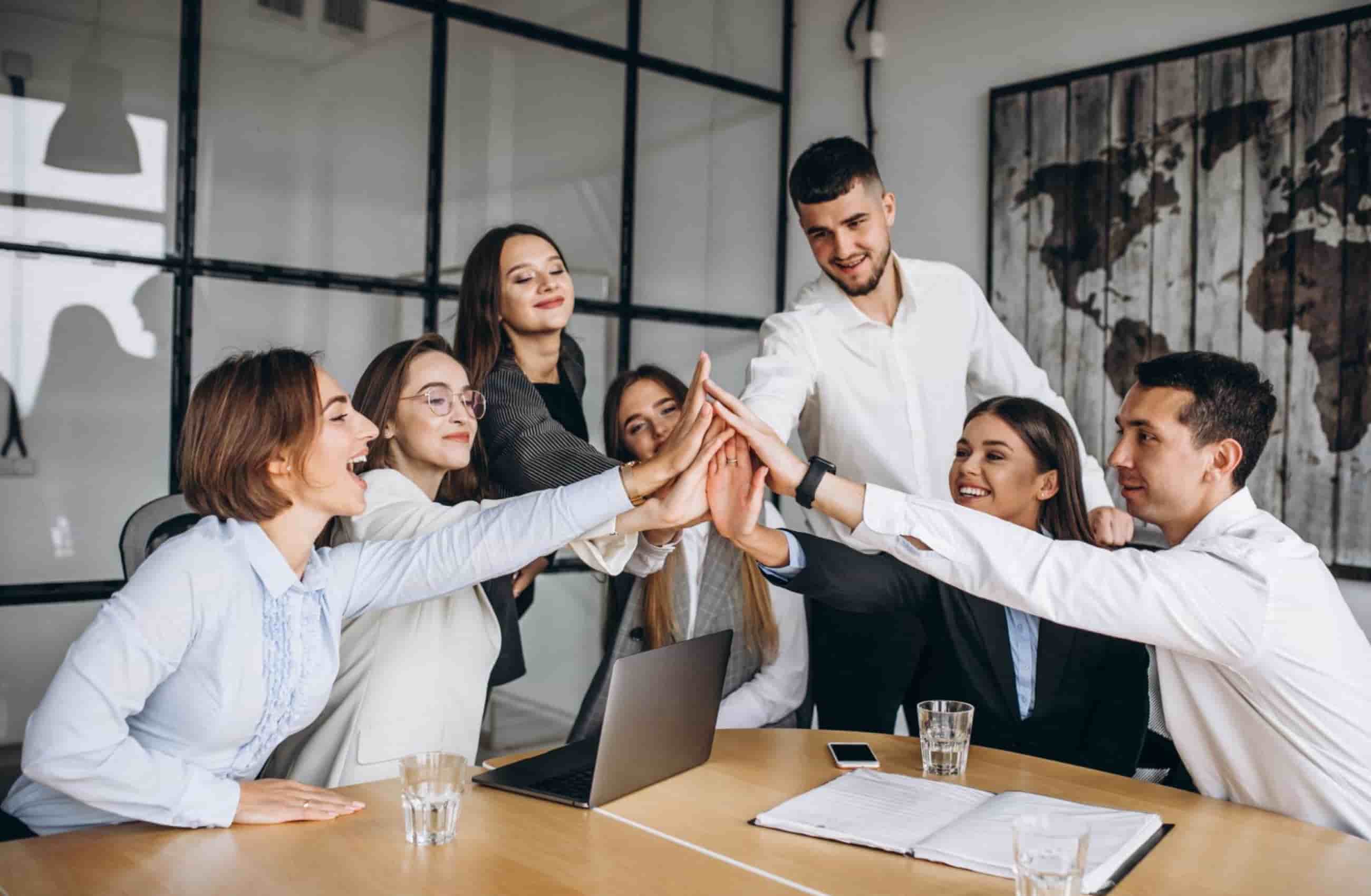 Um grupo corporativo diversificado está reunido ao redor de uma mesa de escritório, unindo as mãos ao centro em um gesto de celebração e trabalho em equipe.&nbsp;