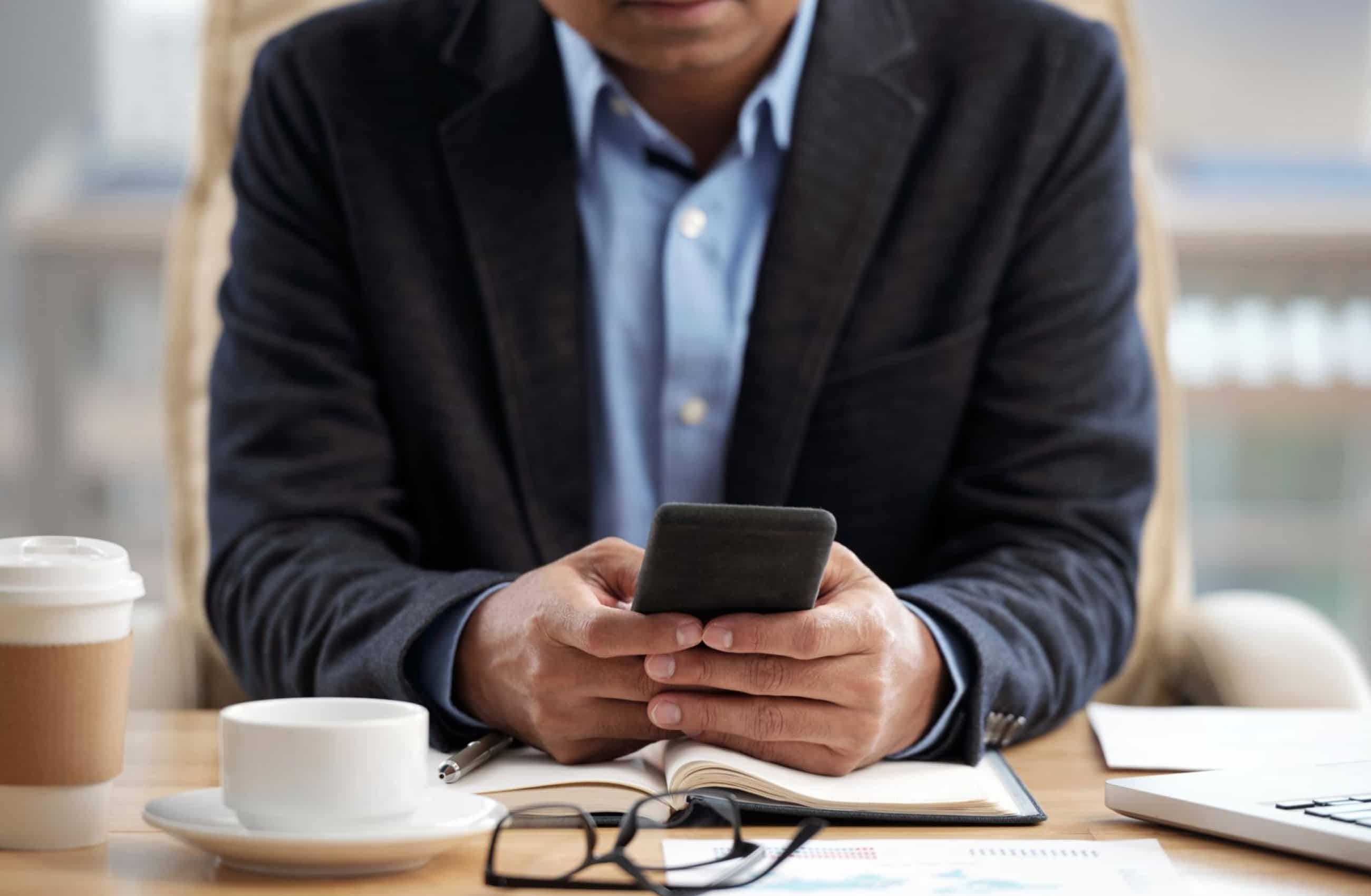 Um executivo de terno escuro segura e utiliza um smartphone com as duas mãos enquanto está sentado à mesa. Na mesa de trabalho, observam-se óculos de leitura e uma xícara de café. 