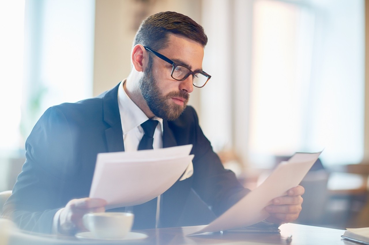 Homem com expressão concentrada, óculos de grau e terno preto, segurando certidão de FGTS nas mãos.