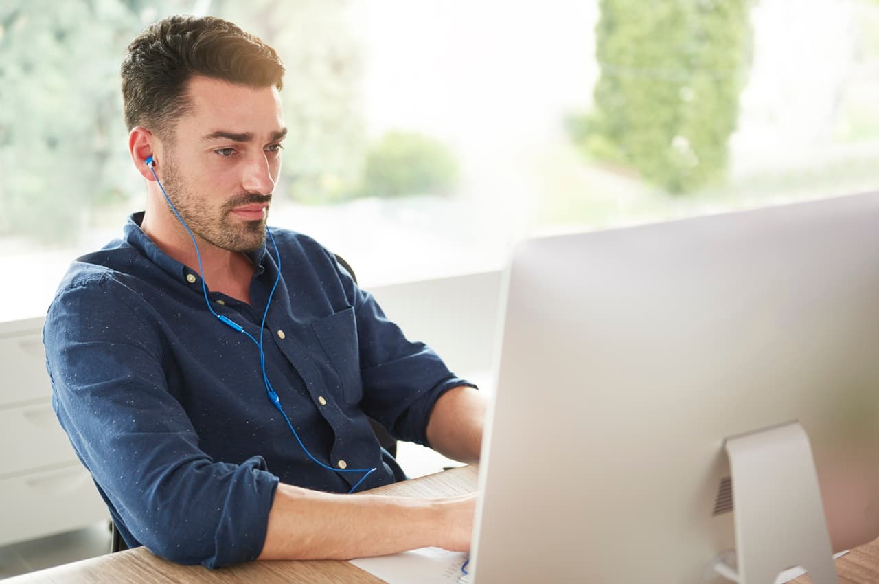 Homem de camiseta azul usando fones de ouvido com fio enquanto trabalha concentrado em um notebook.