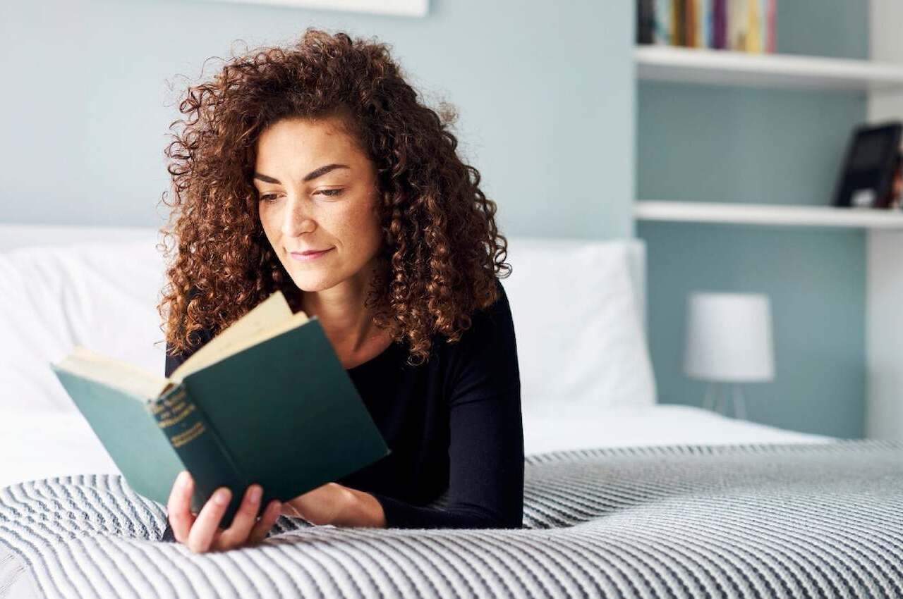 Mulher sorridente de cabelos cacheados, deitada de barriga para baixo na cama, enquanto lê um livro de capa verde.