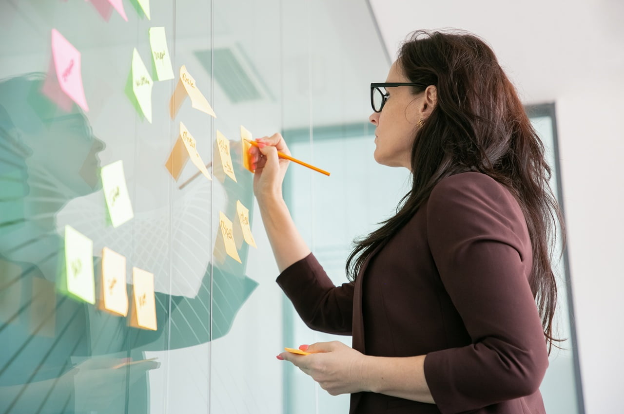 Mulher de óculos escrevendo em post-its coloridos colados em uma parede de vidro.