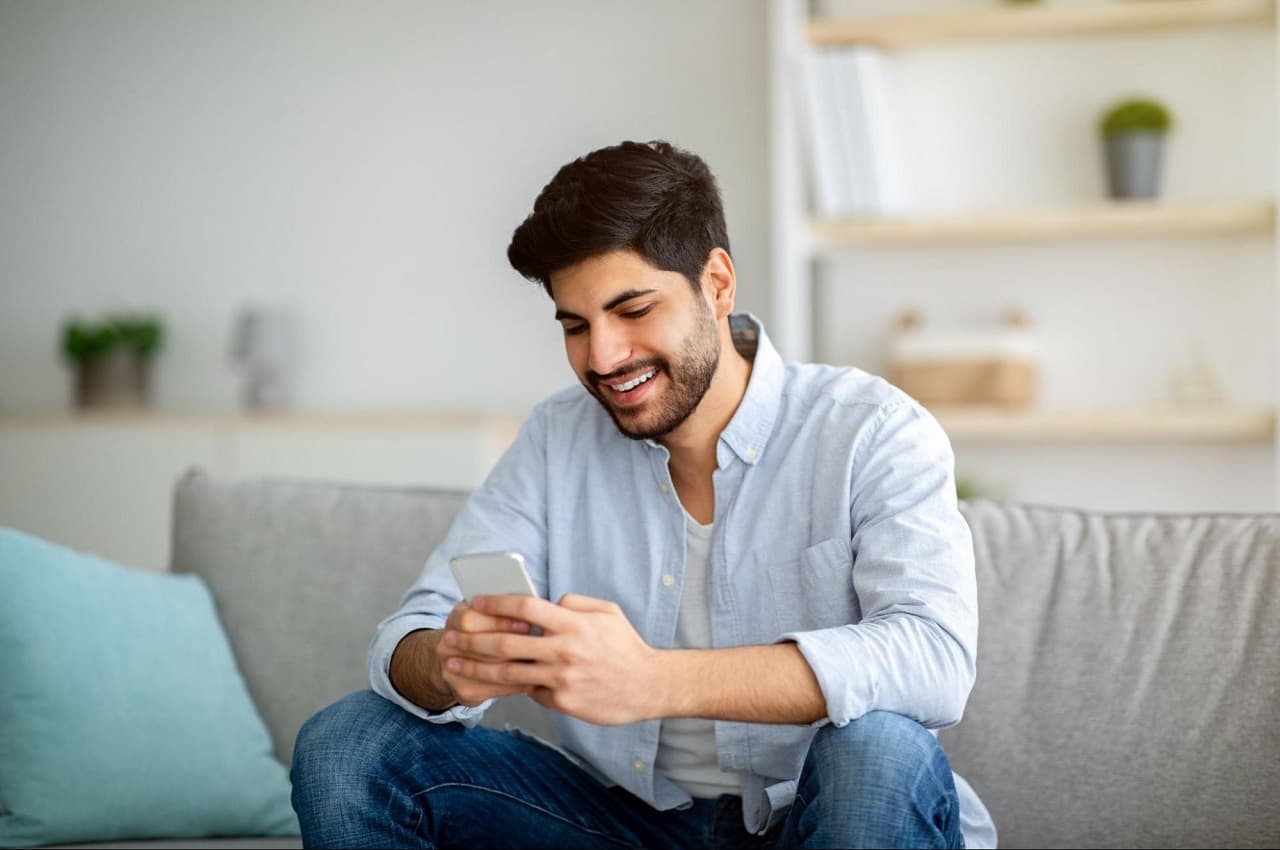 Homem sorridente de cabelos castanhos curtos, sentado no sofá e acessando saque-aniversário FGTS pelo celular.