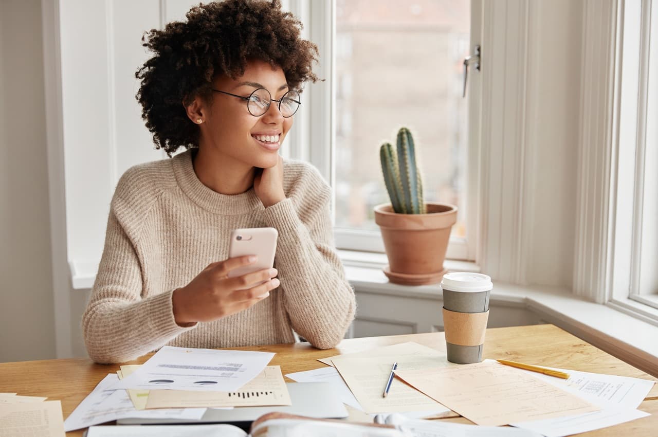 Mulher jovem, de óculos e cabelo cacheado, sorrindo com um celular na mão. Ela está sentada em uma mesa de trabalho com papéis e café.