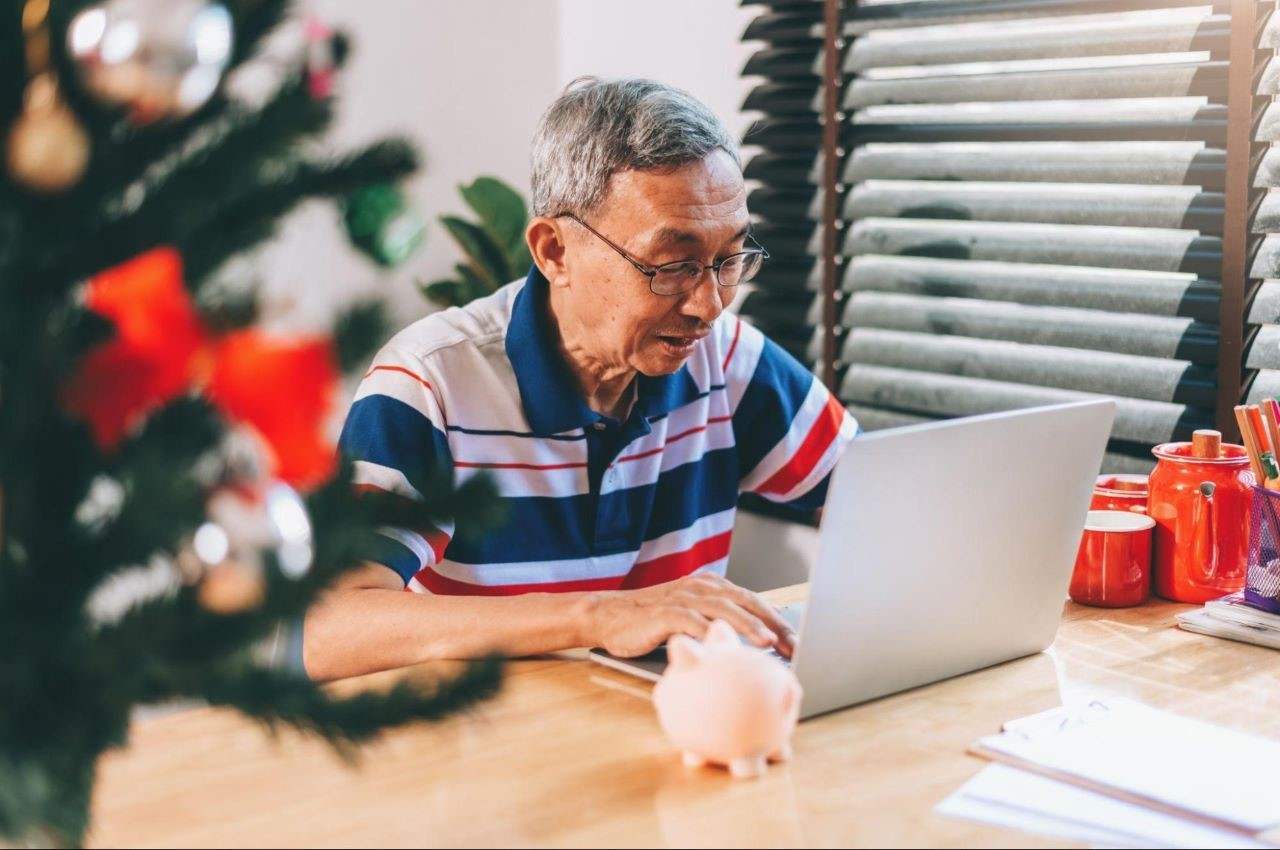 Homem sênior com cabelos grisalhos curtos e óculos de grau, acessando seu computador para consultar FGTS inativo. Em cima da mesa, tem um cofrinho e canecas vermelhas. O homem veste camisa polo branca, azul e vermelha.