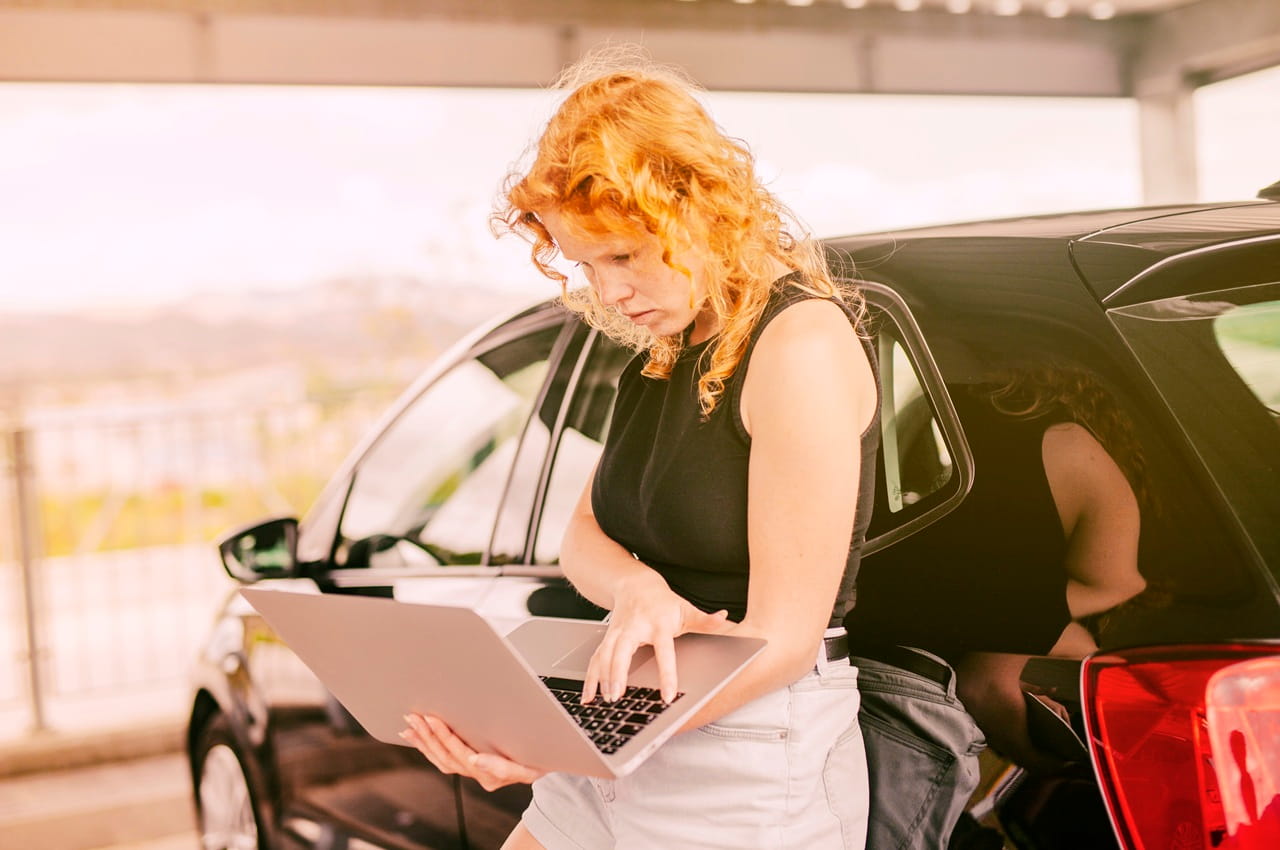 Mulher ruiva pesquisando sobre licenciamento atrasado em seu notebook. Ela está ao lado de um carro preto estacionado.