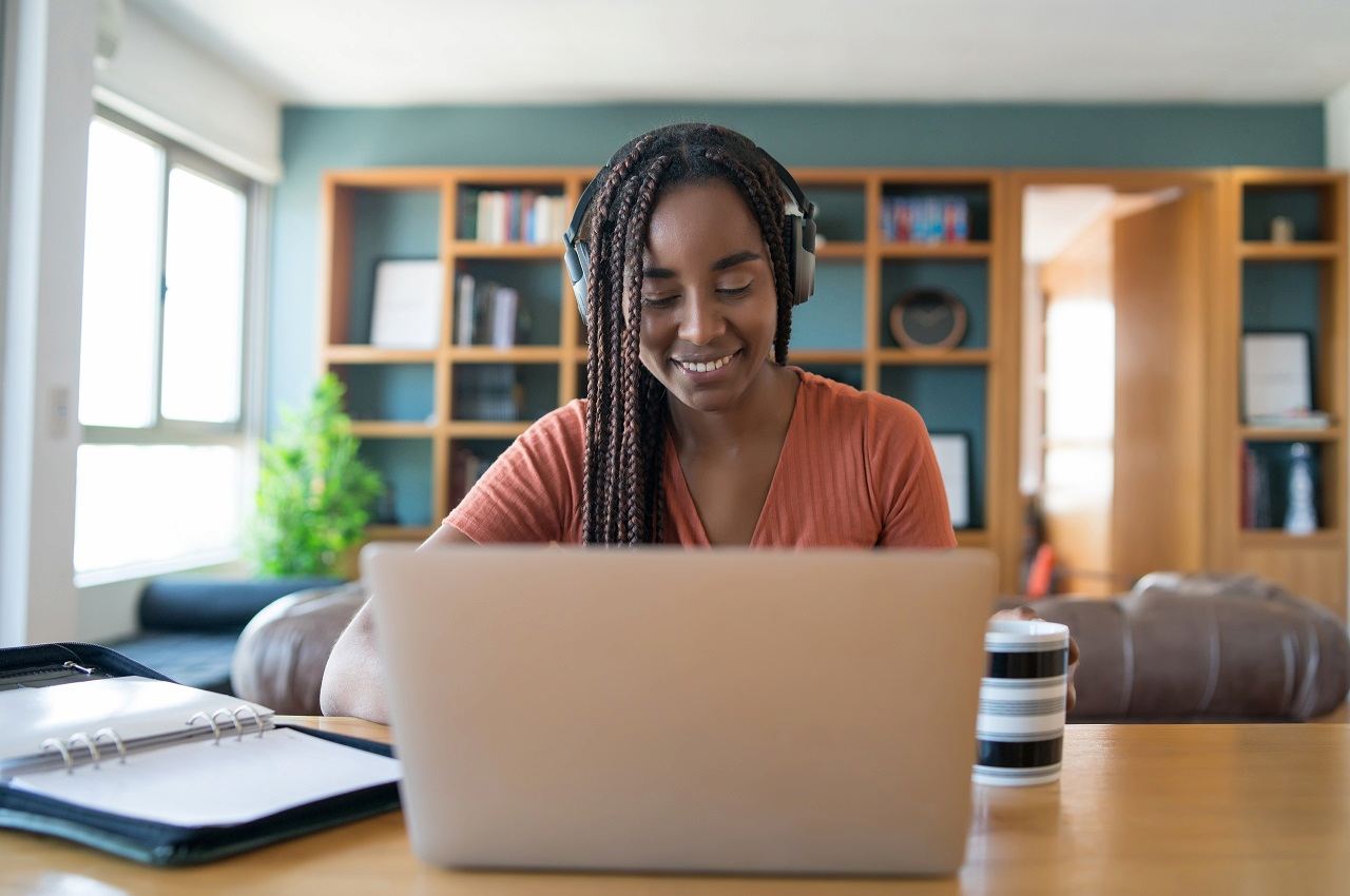 Mulher sorridente com tranças longas, utilizando fones de ouvido bluetooth, fichário e um computador para declarar FGTS no imposto de renda. 