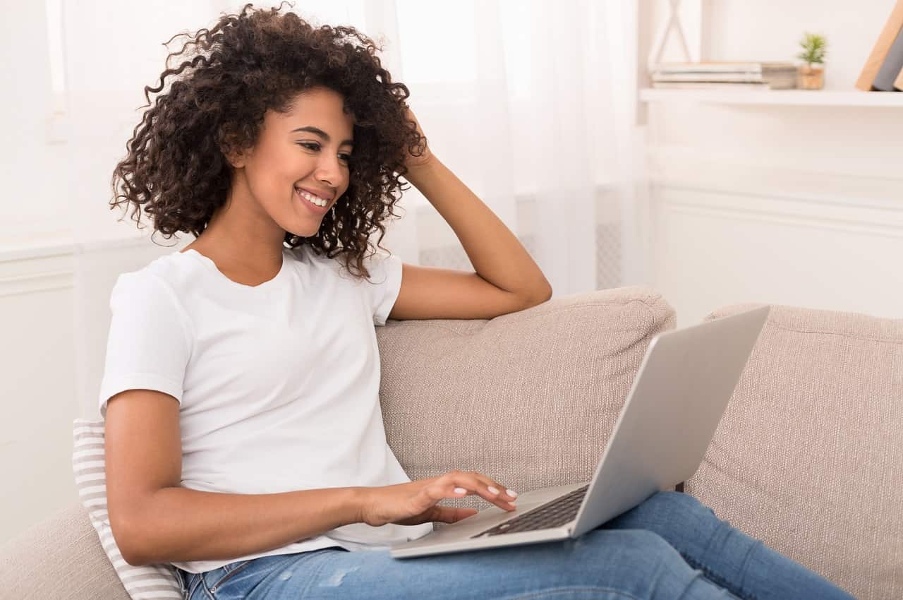 Mulher sorridente vestindo blusa básica branca, calça jeans e sentada no sofá, enquanto utiliza um em seu colo.