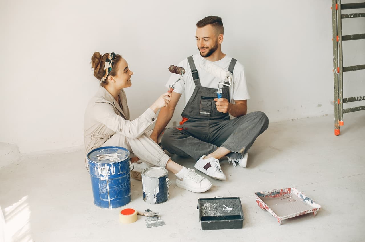 Casal feliz sentado no chão, segurando rolos de pintura. Eles estão cercados por latas de tinta e materiais de pintura, em um cômodo que está passando por reforma.