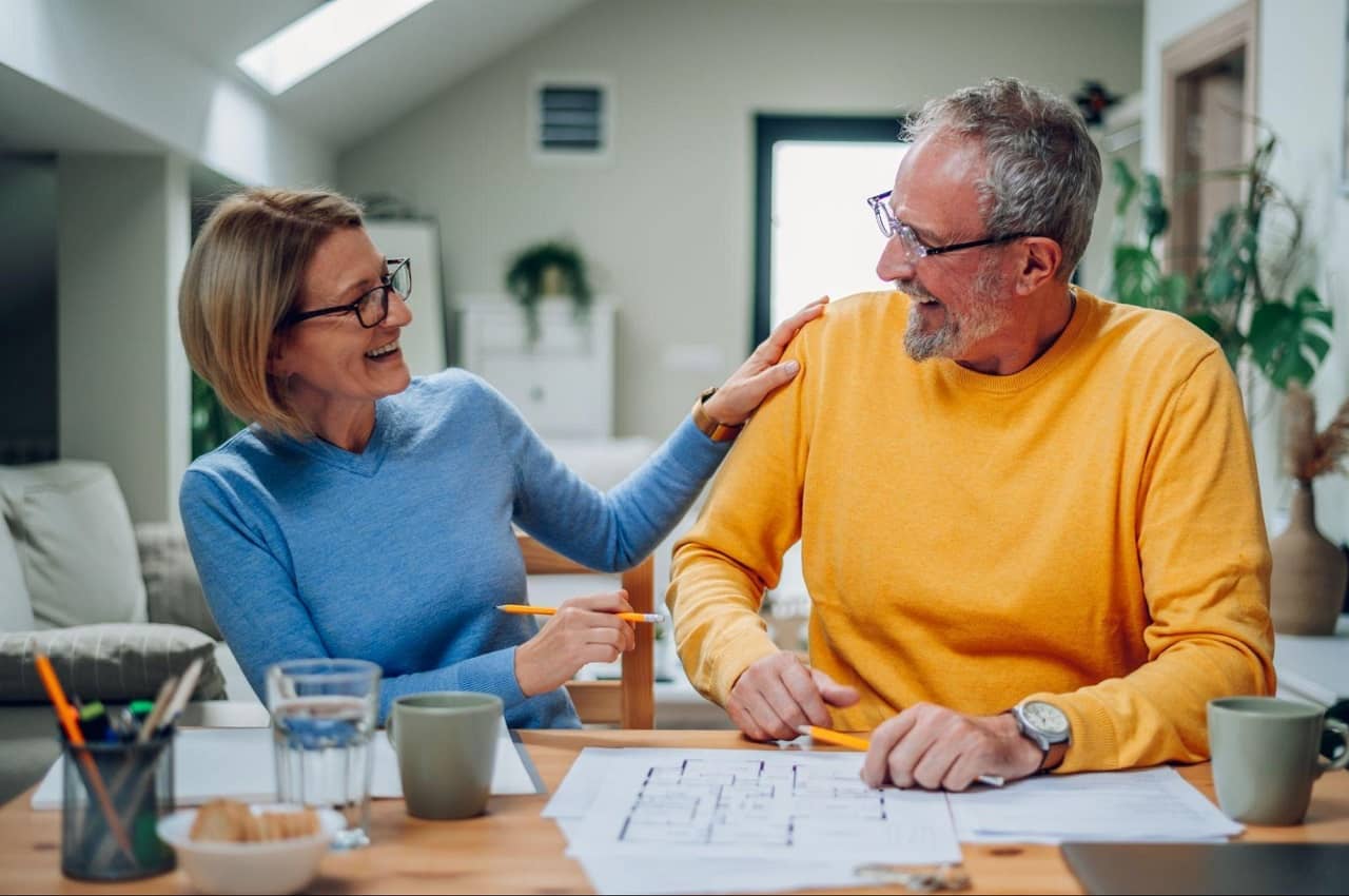 Casal sênior de cabelos grisalhos sorrindo um para o outro, enquanto analisam papéis em cima de uma mesa. Ambos usam moletons azul e amarelo.