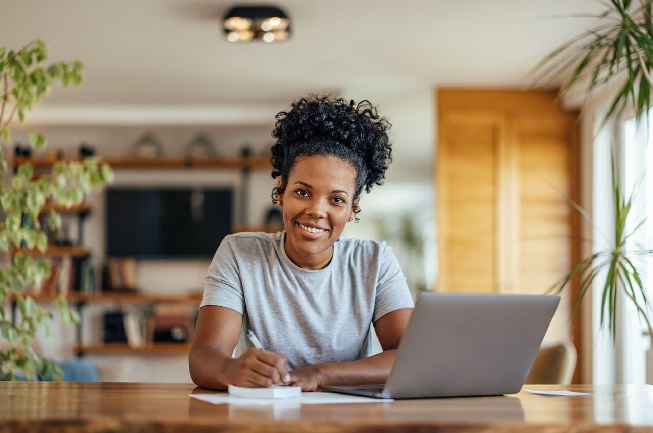 Mulher sorridente utilizando computador e anotações em bloco de notas. Ela veste blusa básica cinza e está em seu escritório.