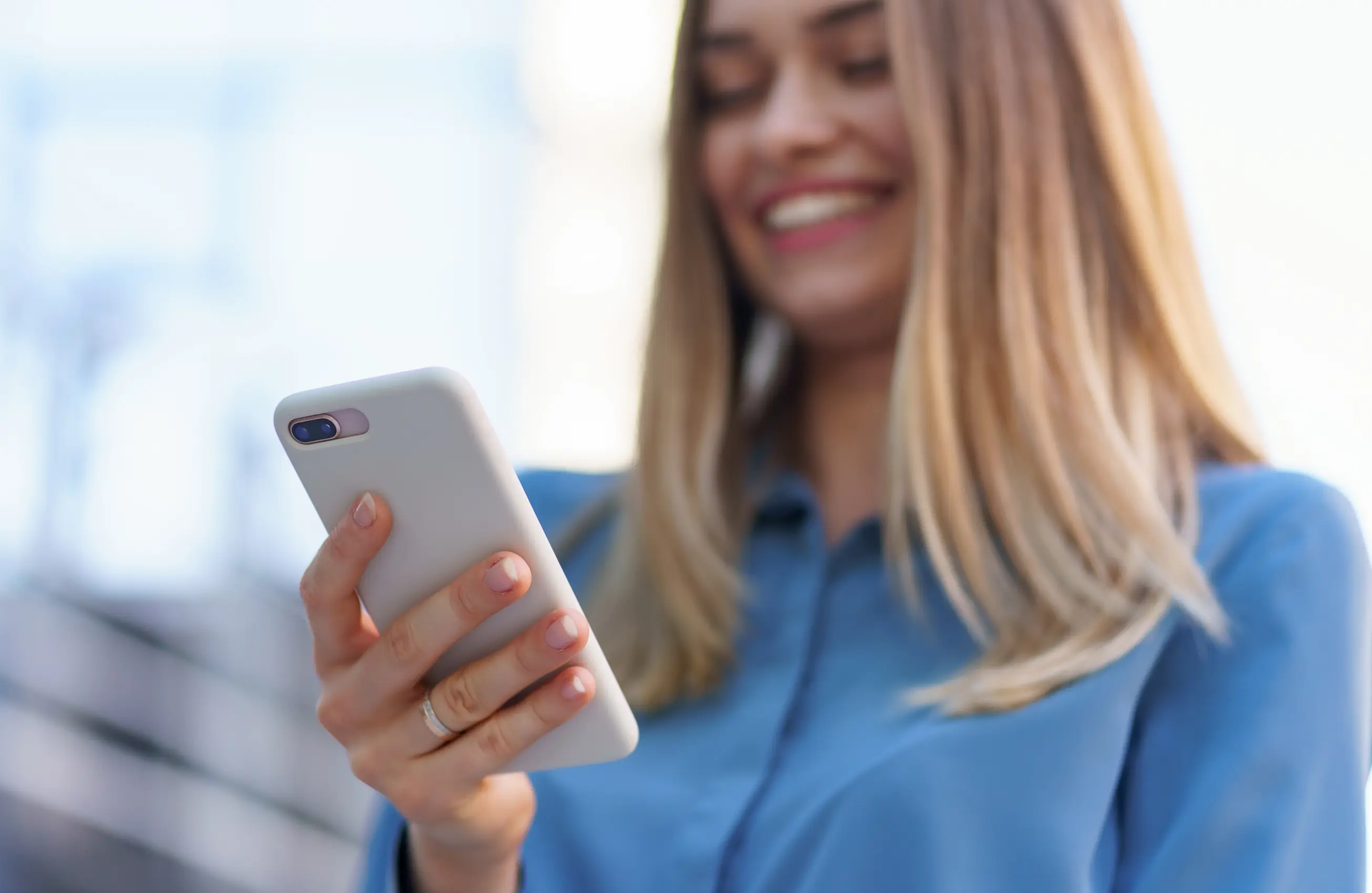 Uma mulher sorrindo enquanto utiliza um smartphone. Seu rosto está parcialmente desfocado.