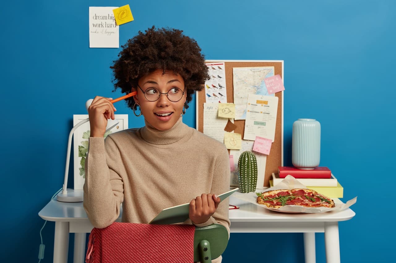 Mulher sentada em uma cadeira enquanto segura um caderno de anotações em uma das mãos e um lápis na outra. Ela está com uma expressão de descoberta.