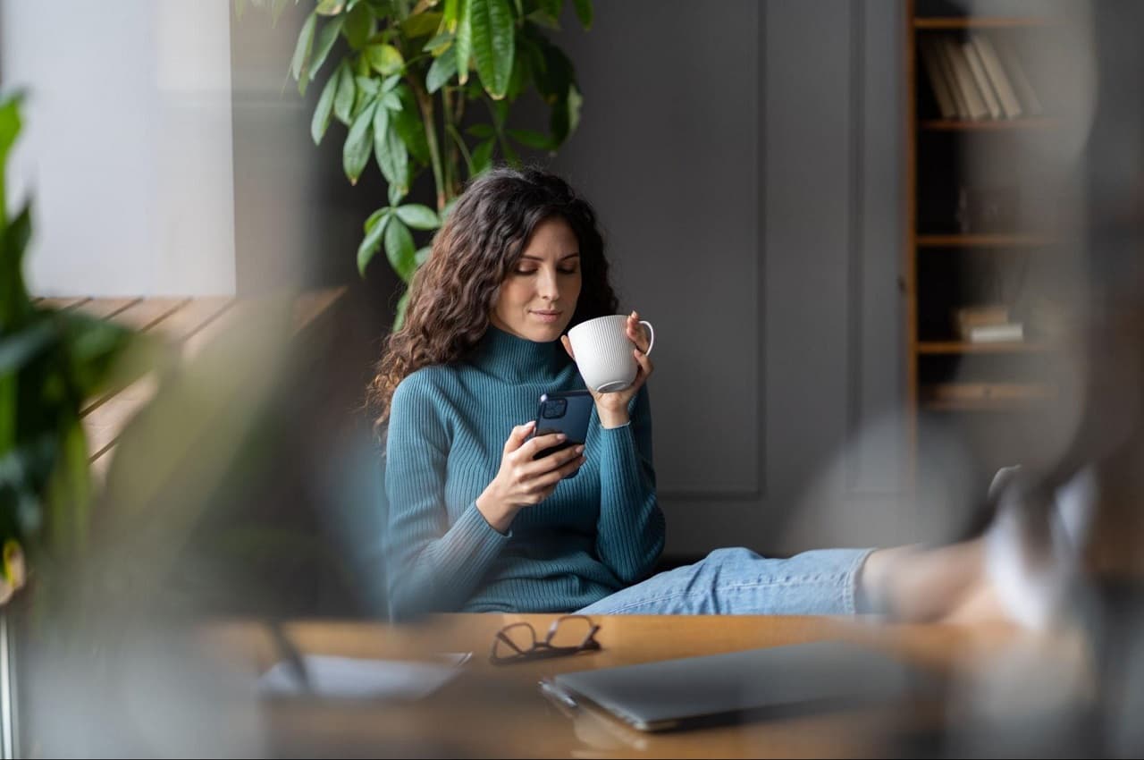 Mulher sorridente no ambiente de trabalho, segurando xícara de café e utilizando celular para antecipar saque-aniversário. Ela apoia os pés na mesa do escritório e inclina as costas na cadeira.