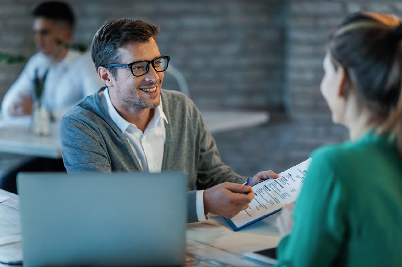 Homem de óculos sorrindo enquanto segura um documento e conversa com uma mulher sentada à sua frente, em uma mesa com notebook.