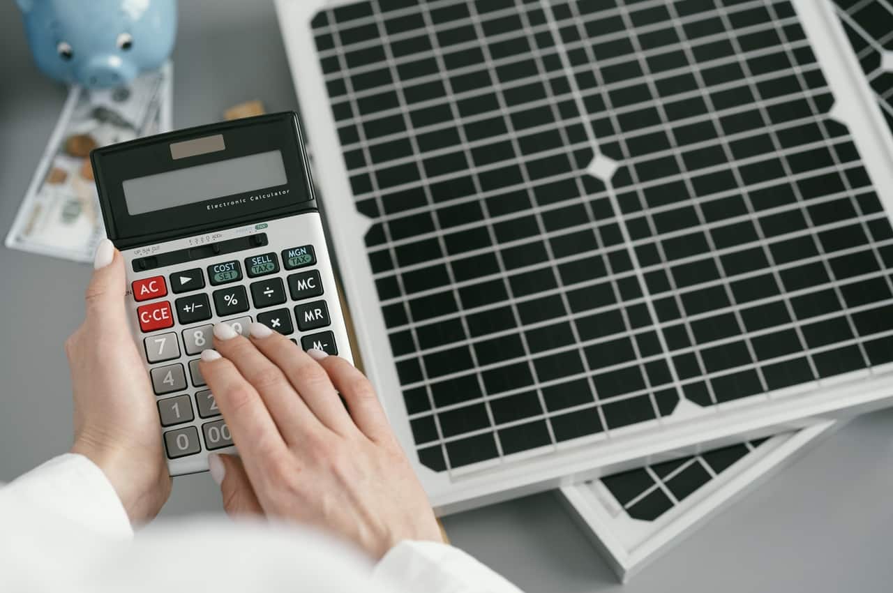 Mãos usando uma calculadora ao lado de um pequeno painel solar e notas de dinheiro, simbolizando a taxação do sol.