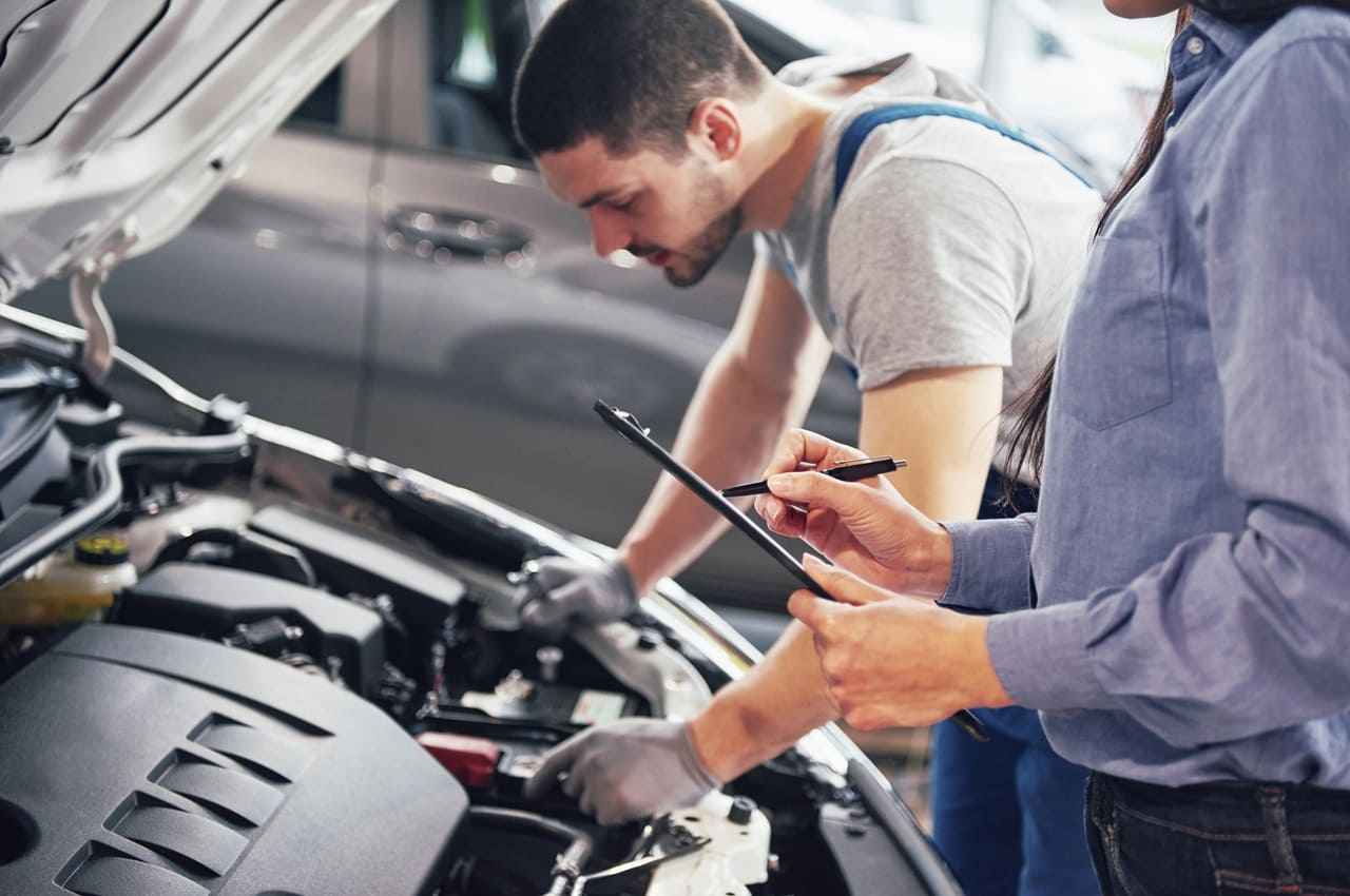 Homem verificando o motor de um carro enquanto uma mulher, ao seu lado, faz anotações em um tablet. Eles estão no processo de vistoria cautelar.