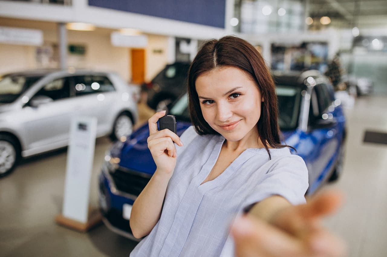 Mulher sorridente segurando a chave de um carro em frente a um veículo azul, em uma concessionária.