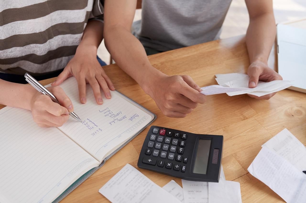 Duas pessoas (seus rostos não aparecem na imagem) realizando anotações de contas e utilizando calculadora.