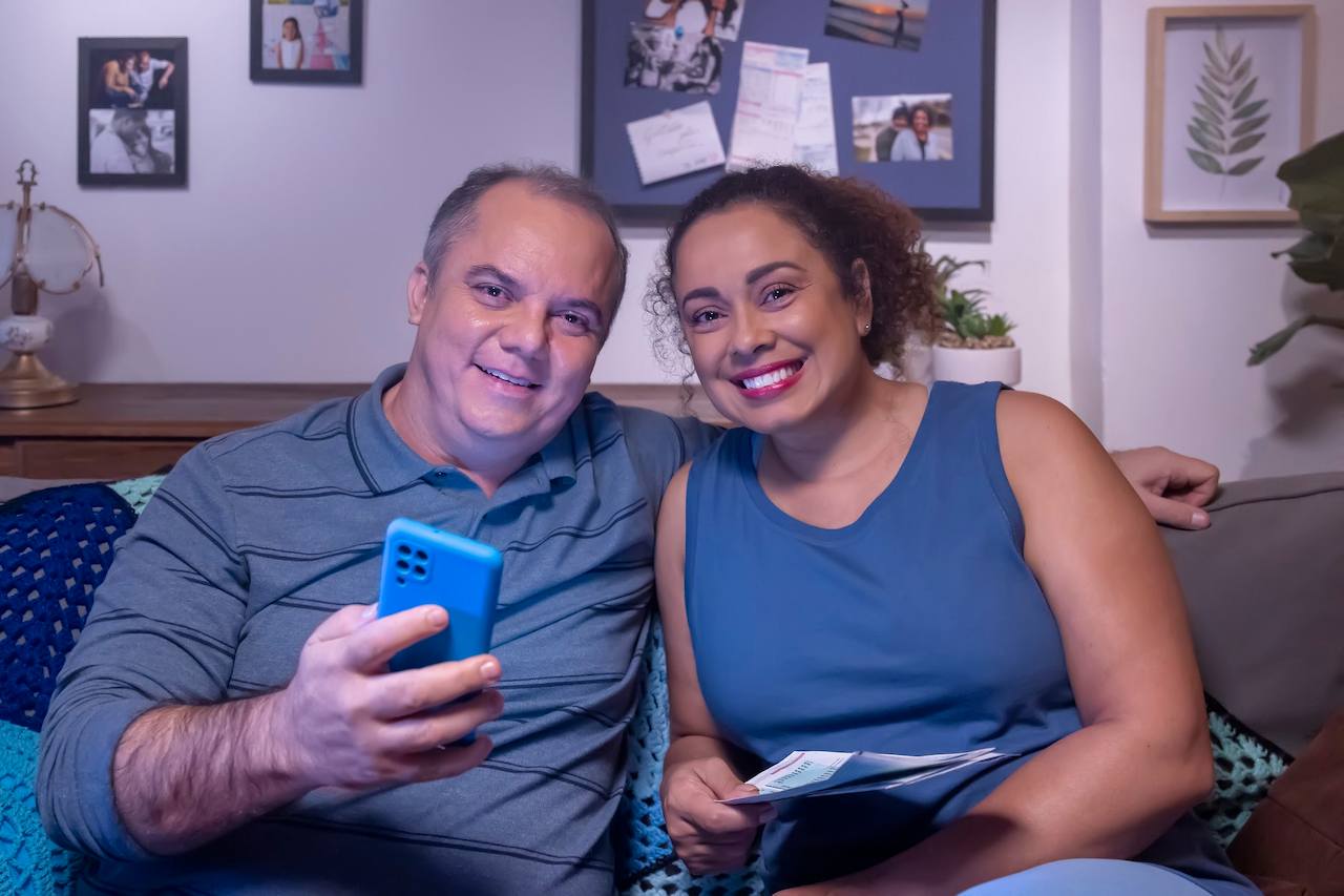 Casal sorridente sentado no sofá de sua casa acessando o celular e vendo contas a pagar. O homem está abraçando a mulher, enquanto utiliza o celular para acessar empréstimo FGTS.