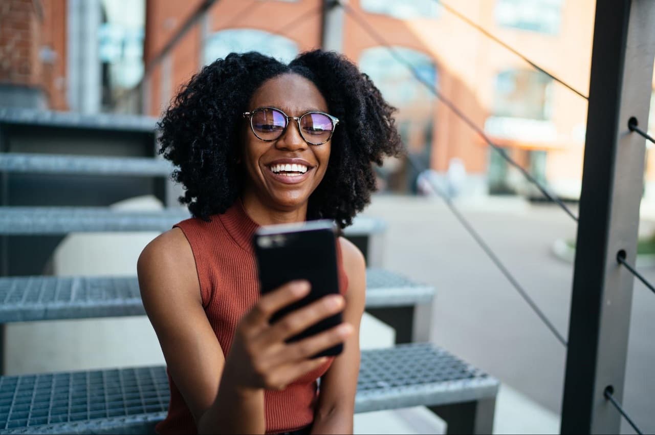 Mulher sorridente com óculos de grau, sentada na escada de um prédio e acessando saque-aniversário FGTS pelo celular. Ela veste regata marrom e calça jeans preta.