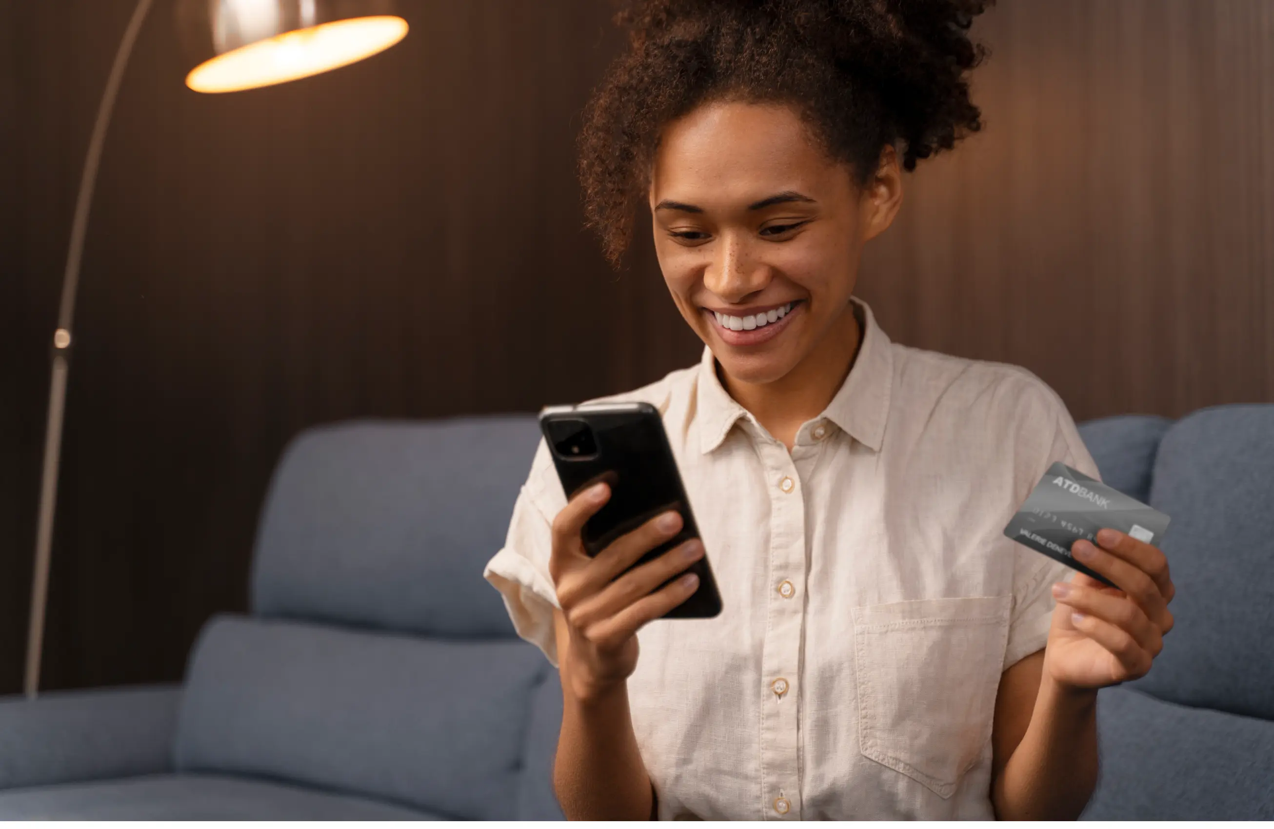 Mulher negra sorrindo em ambiente interno, enquanto segura um cartão e um celular.
