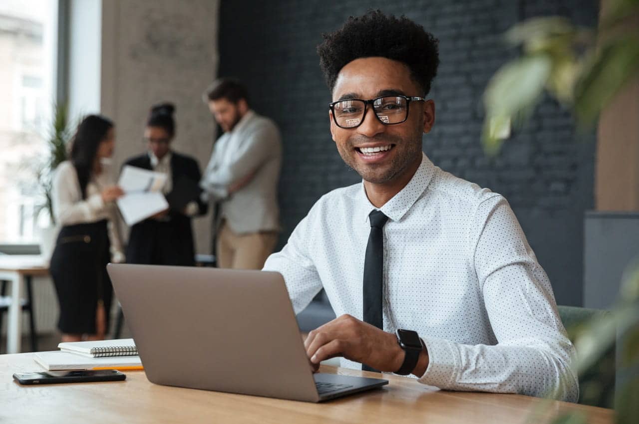 Homem de camisa branca com gravata preta sorrindo, enquanto usa um notebook em uma mesa de escritório. Ao fundo, há pessoas conversando e analisando documentos.