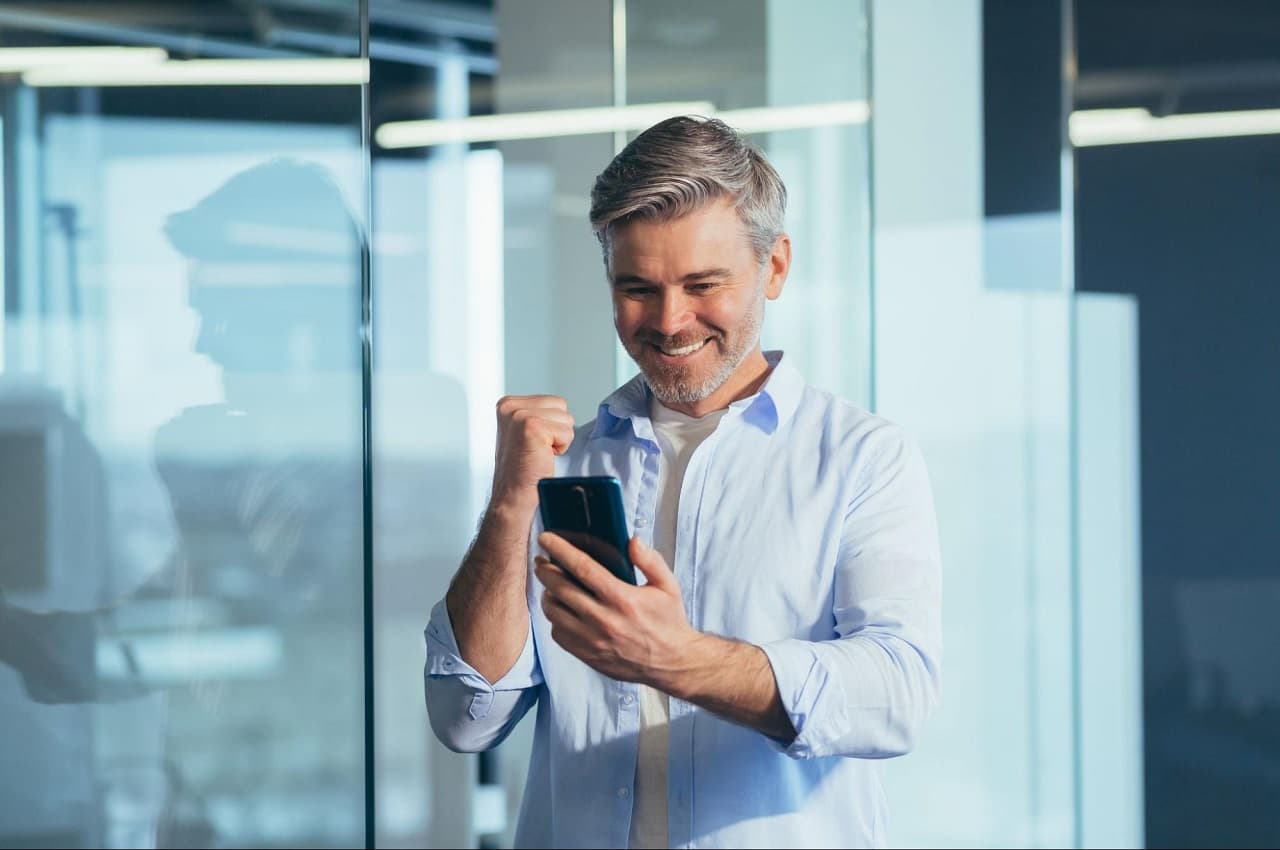 Homem de cabelos grisalhos com expressão sorridente, segurando celular e realizando gesto comemorativo com as mãos.