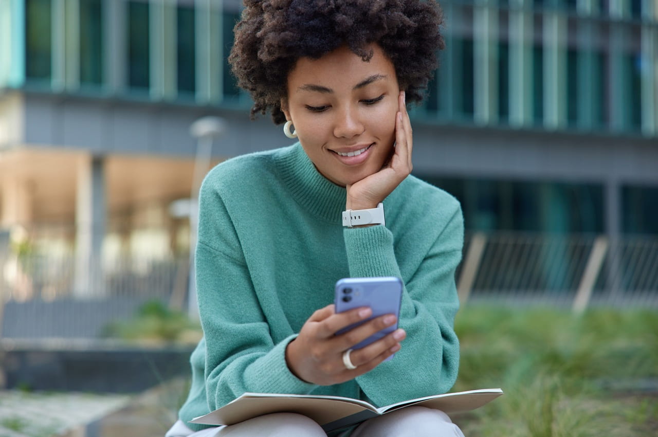 Mulher de cabelo cacheado olhando para o celular com um semblante sorridente. Ela está ao ar livre e pesquisando sobre o Simulador de Empréstimo com Garantia de Veículo.