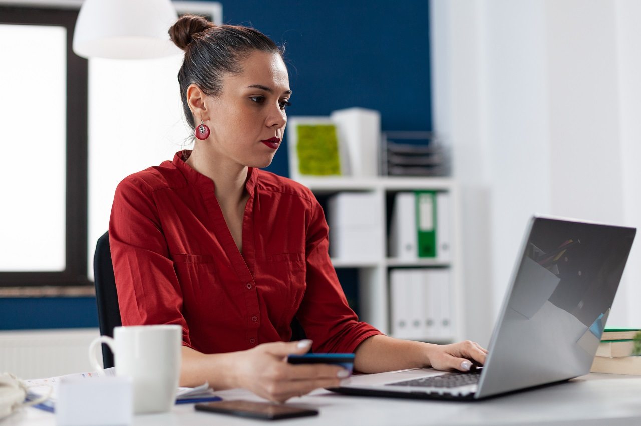 Mulher com expressão concentrada, vestindo blusa social vermelha e utilizando computador no seu ambiente de trabalho. Ela segura um cartão azul.