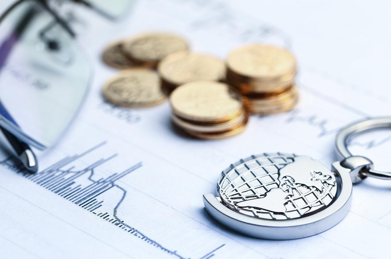 Pilhas de moedas dispostas sobre documentos financeiros. Há um chaveiro do globo terrestre ao lado, simbolizando operações de trade finance.