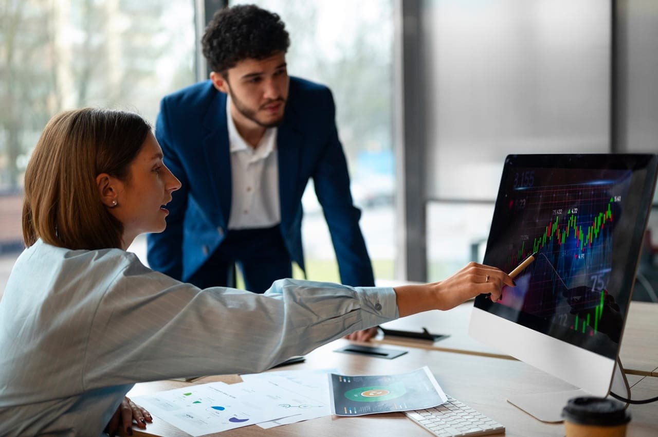 Duas pessoas, no interior de um escritório, observando um monitor com gráficos, como um sinal de aprendizado sobre Letras Financeiras.