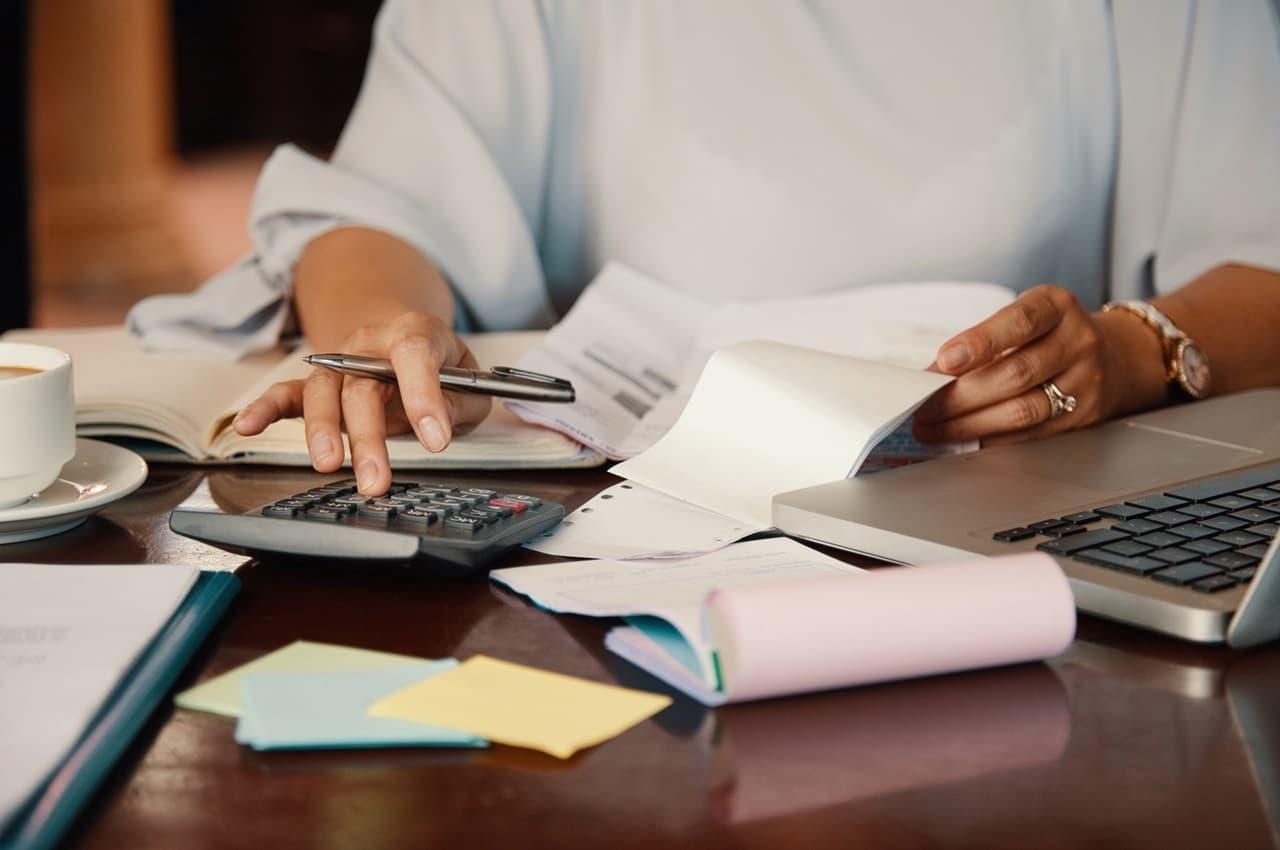 Pessoa usando calculadora e um notebook para calcular como liberar margem consignável. Na mesa em que ela está apoiada, há post-its, documentos e extratos bancários.
