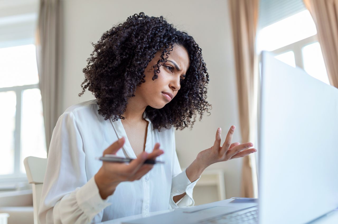 Mulher com expressão concentrada segurando uma caneta prata, com as mãos levantadas em direção a tela do computador. Ela veste blusa social branca e está no escritório.
