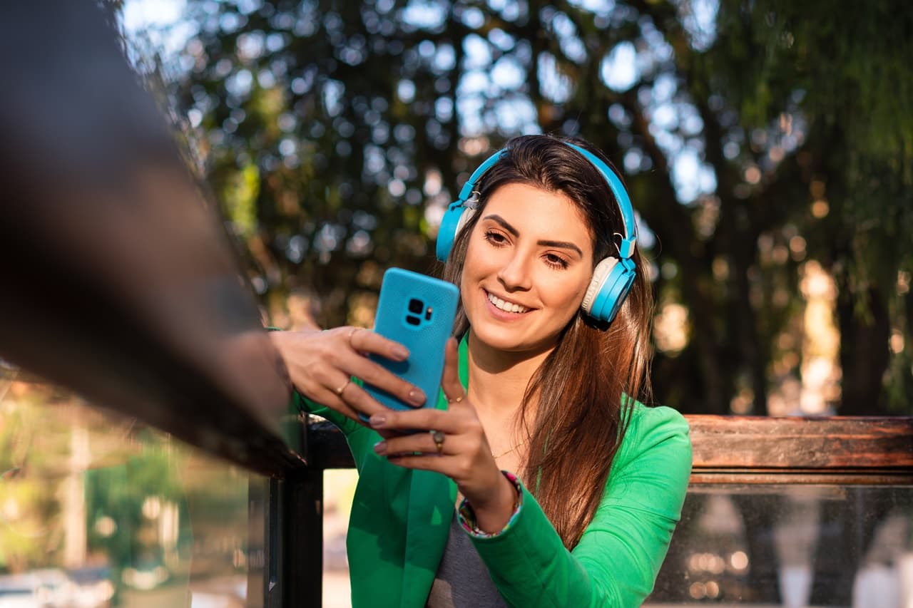 Mulher sorrindo com fone de ouvido, no parque, com celular na mão