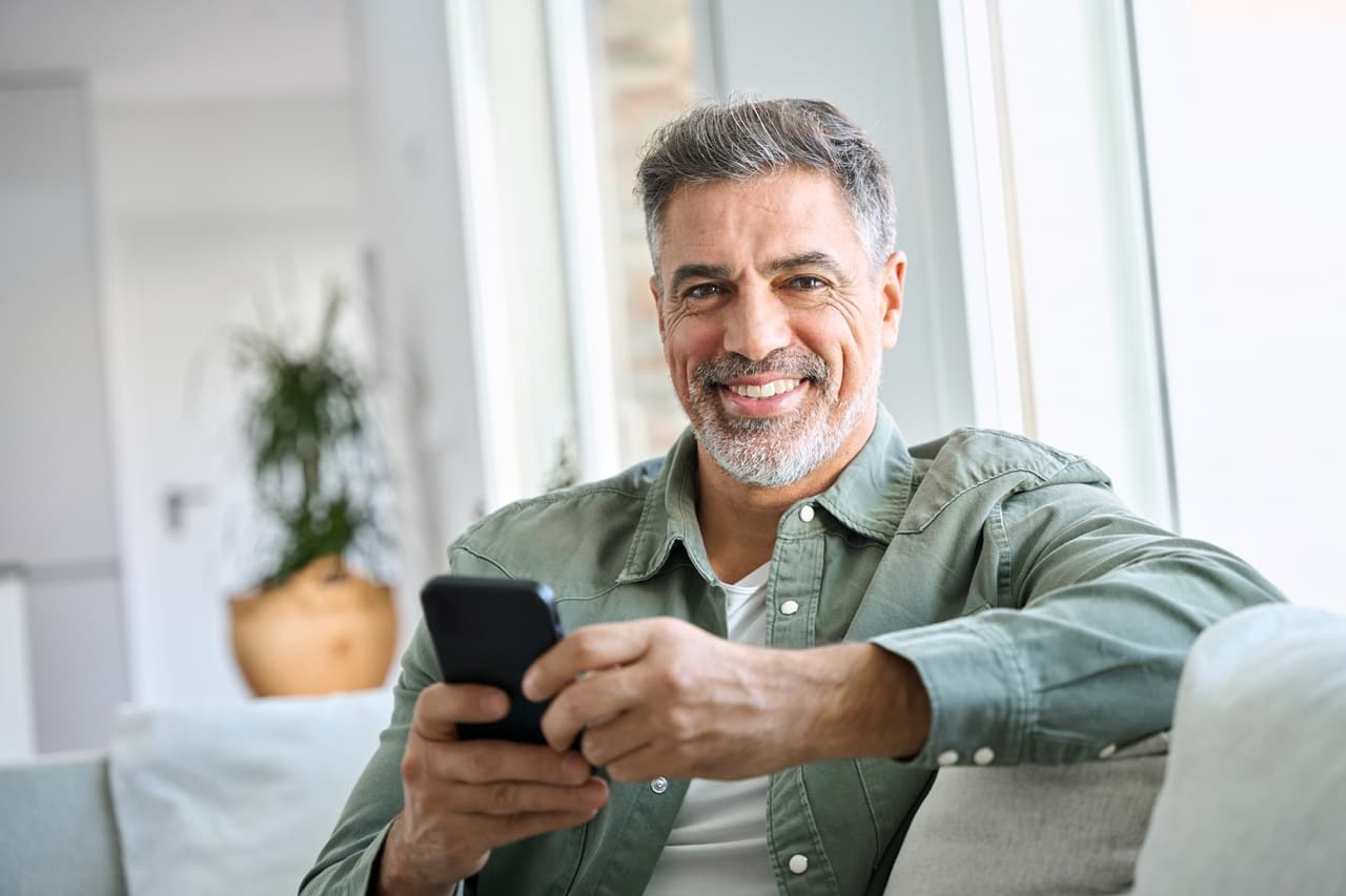 homem sentado em sofá sorrindo, com celular na mão