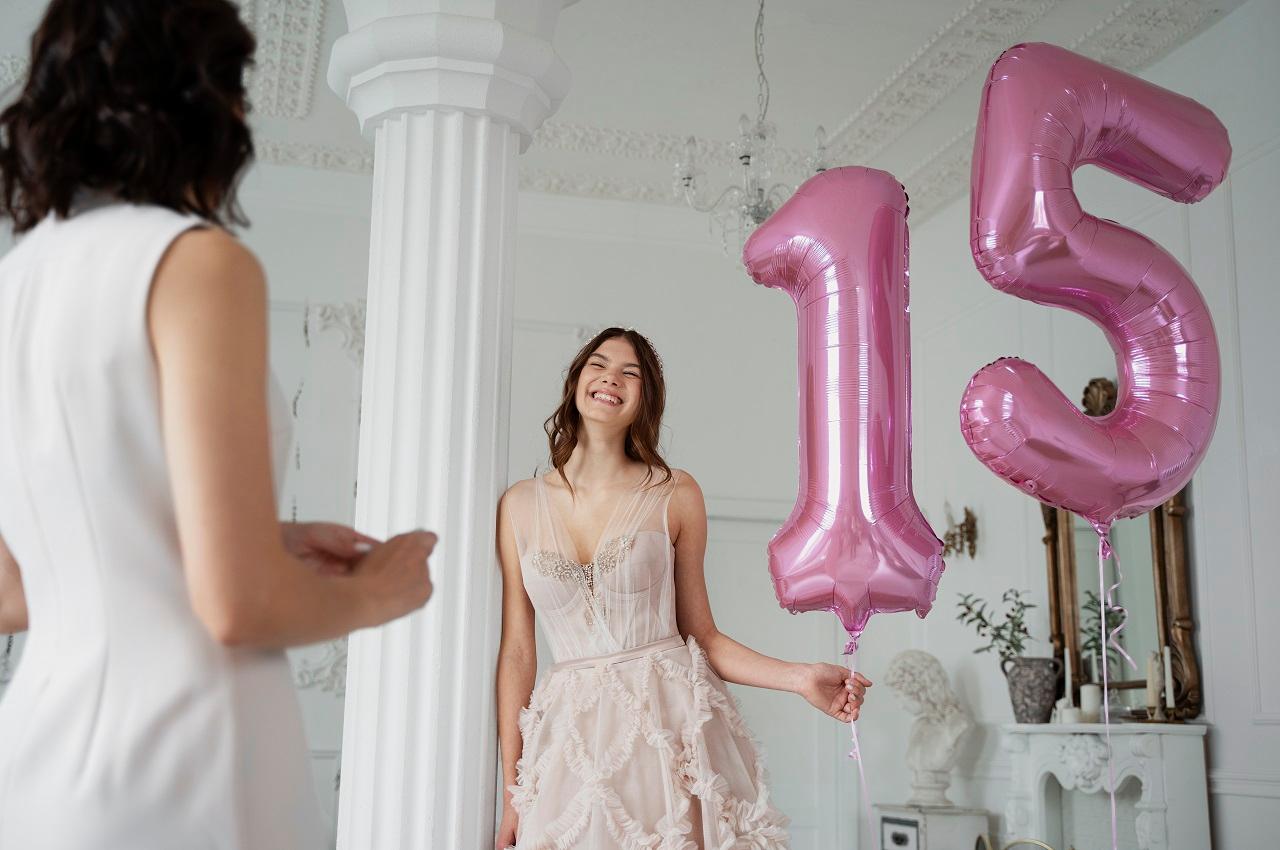 Menina de 15 anos sorrindo para sua mãe e segurando balões com o número 15. Ela veste um vestido rosa claro de festa de 15 anos com brilho no busto e babados na saia.